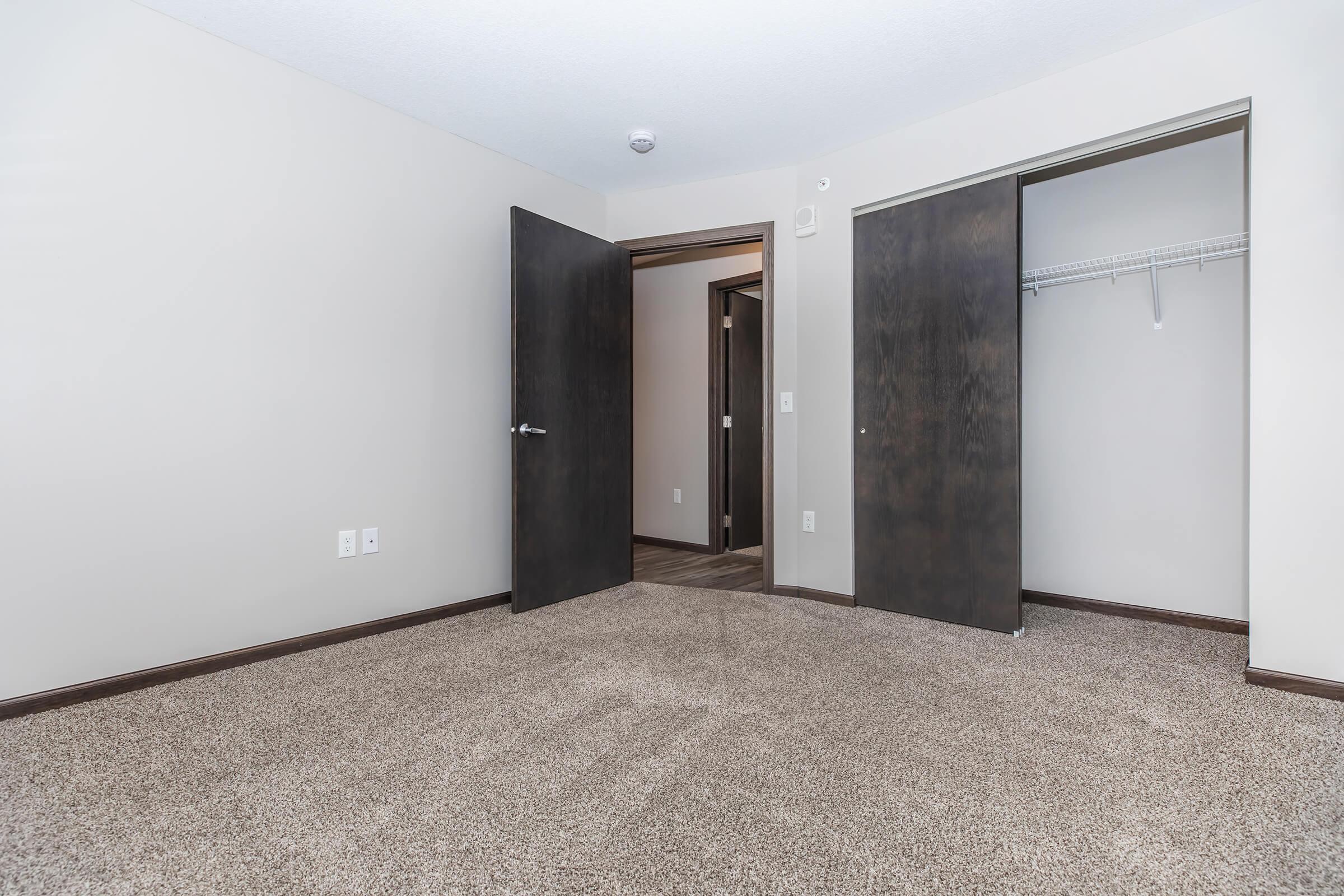 A spacious, empty room with light brown carpet and neutral-colored walls. Two dark wood doors are partially open, leading to another room and a closet with a hanging rod. Natural light filters in from an unseen source, creating a bright and inviting atmosphere.
