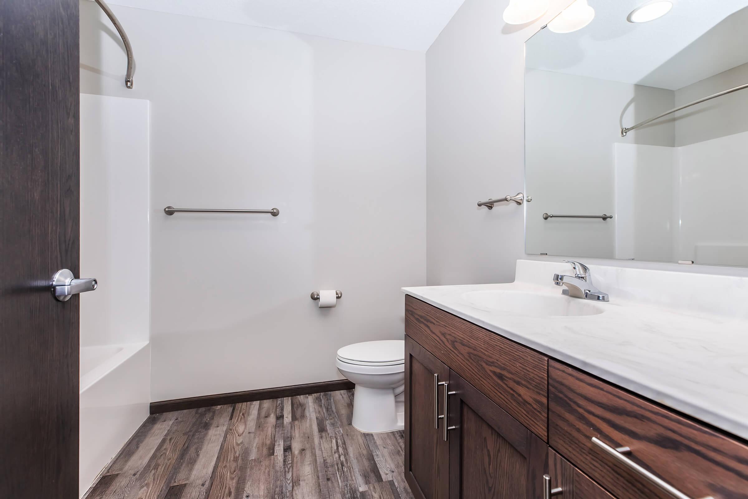 A modern bathroom featuring a white bathtub, a toilet, and a dark wood vanity with a marble countertop. There is a large mirror above the sink, with two light fixtures above. The walls are painted in a neutral tone, and the floor is covered with wood-like vinyl planks.