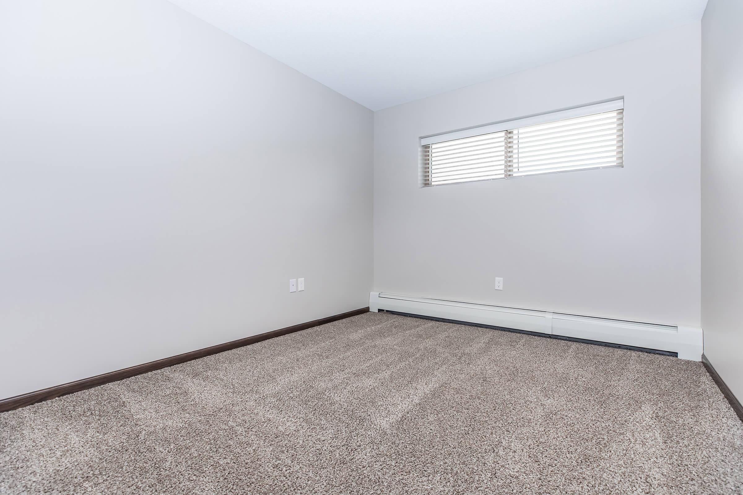 A vacant room with light beige walls, a small window featuring horizontal blinds, and a plush carpet in a similar shade. The floor is clear of furniture, creating a spacious, empty feel. White baseboards line the bottom of the walls, and there are two electrical outlets visible.
