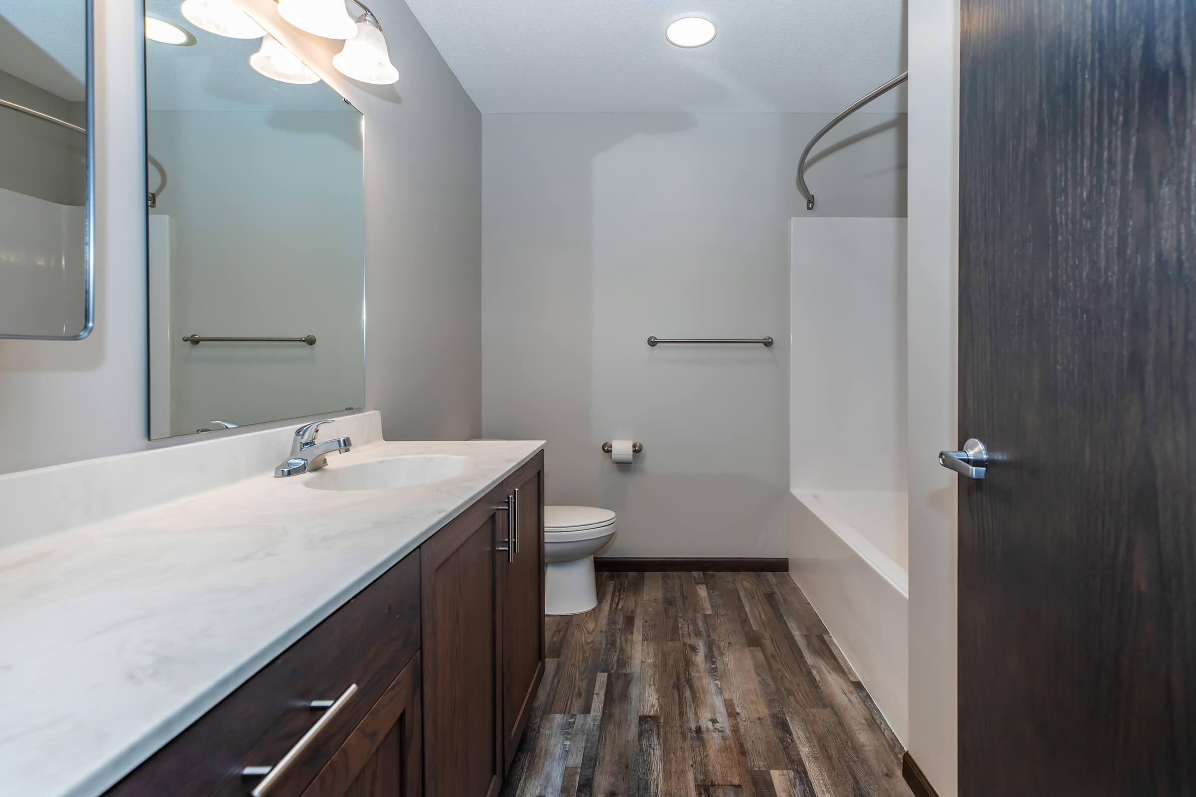 A modern bathroom featuring a marble countertop vanity with a mirror, a white bathtub with a shower, a toilet, and dark wooden cabinetry. The flooring consists of wooden planks, and the walls are a soft gray color. Natural light is provided by overhead lighting.