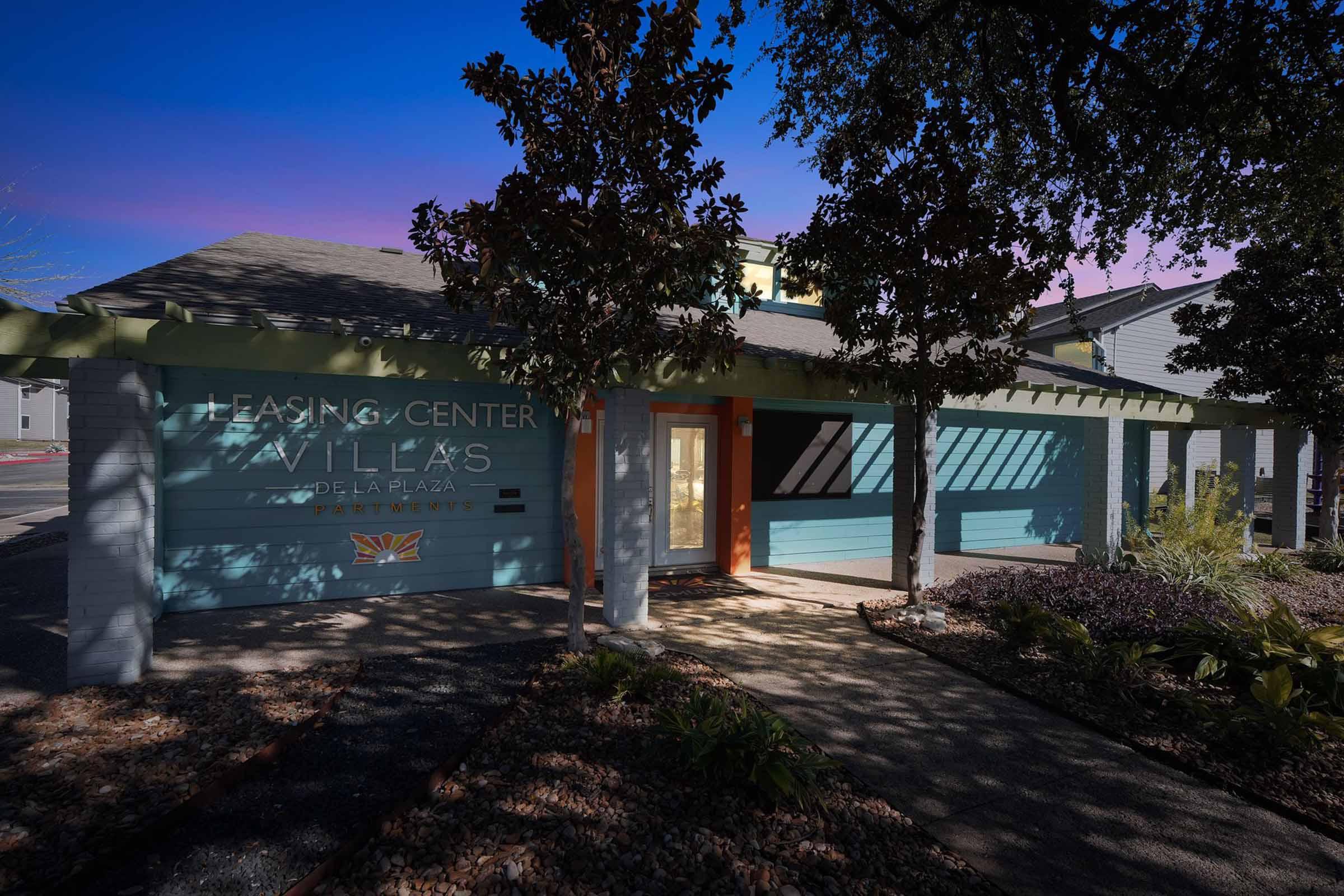 A contemporary leasing center building with a turquoise exterior and orange accents. The entrance features large windows and a welcoming door. Surrounding the building are landscaped areas with plants and pathways, set against a twilight sky.