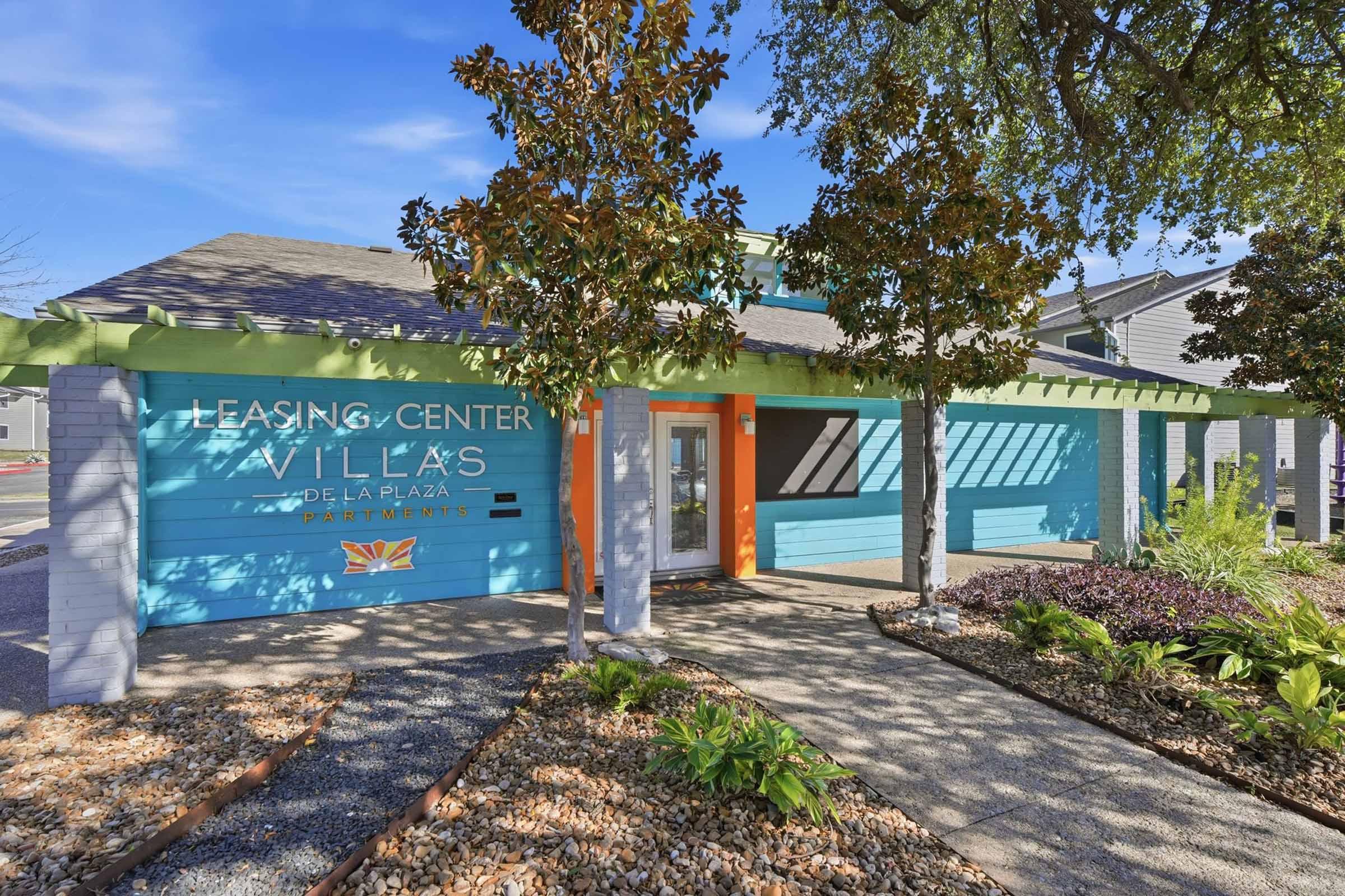 A brightly colored leasing center for "Villas del La Plaza" apartments, featuring a vibrant blue exterior with orange accents and a landscaped entrance. The building is surrounded by greenery and decorative stones, creating an inviting atmosphere.