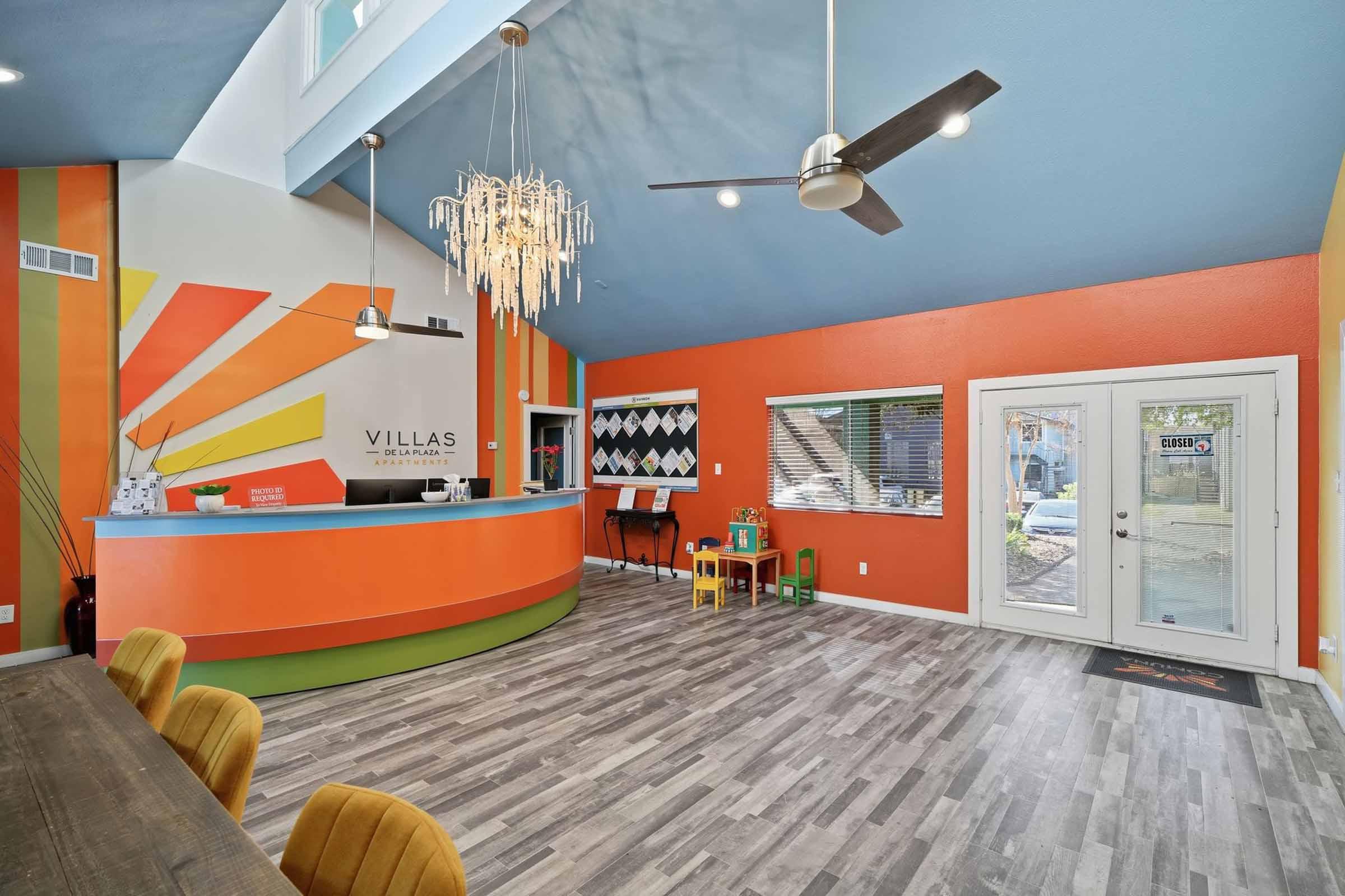 Modern reception area with a vibrant color scheme featuring orange and blue walls. A stylish chandelier hangs from the ceiling. There is a curved reception desk with decorative elements and seating for visitors. Large windows provide natural light, and colorful chairs are placed in the corner of the room.