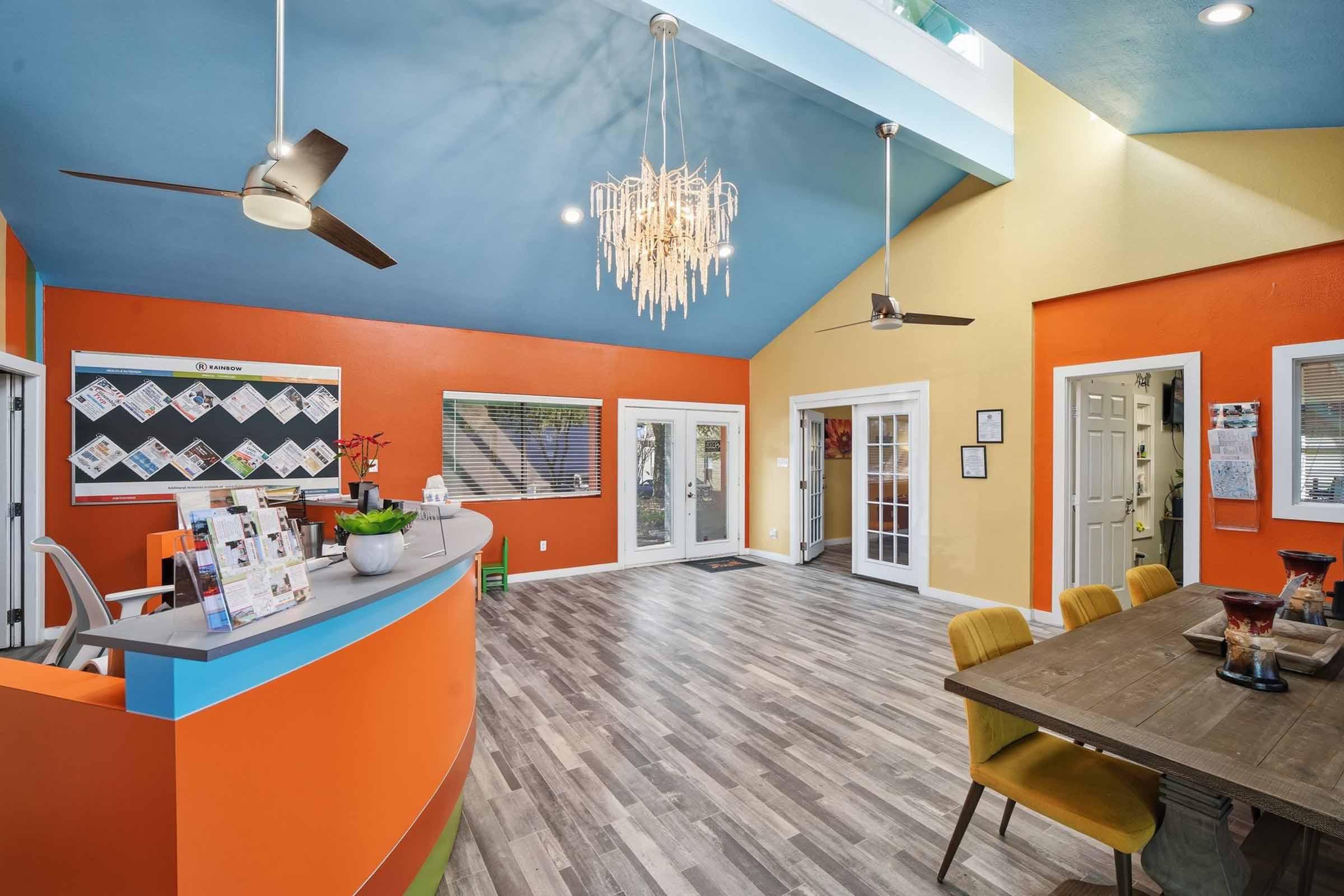 Interior of a brightly colored office reception area featuring a curved front desk, a chandelier, and large windows. The walls are painted in orange and yellow, with a blue ceiling. There are wooden floors and a dining table with chairs, creating a welcoming and modern atmosphere.