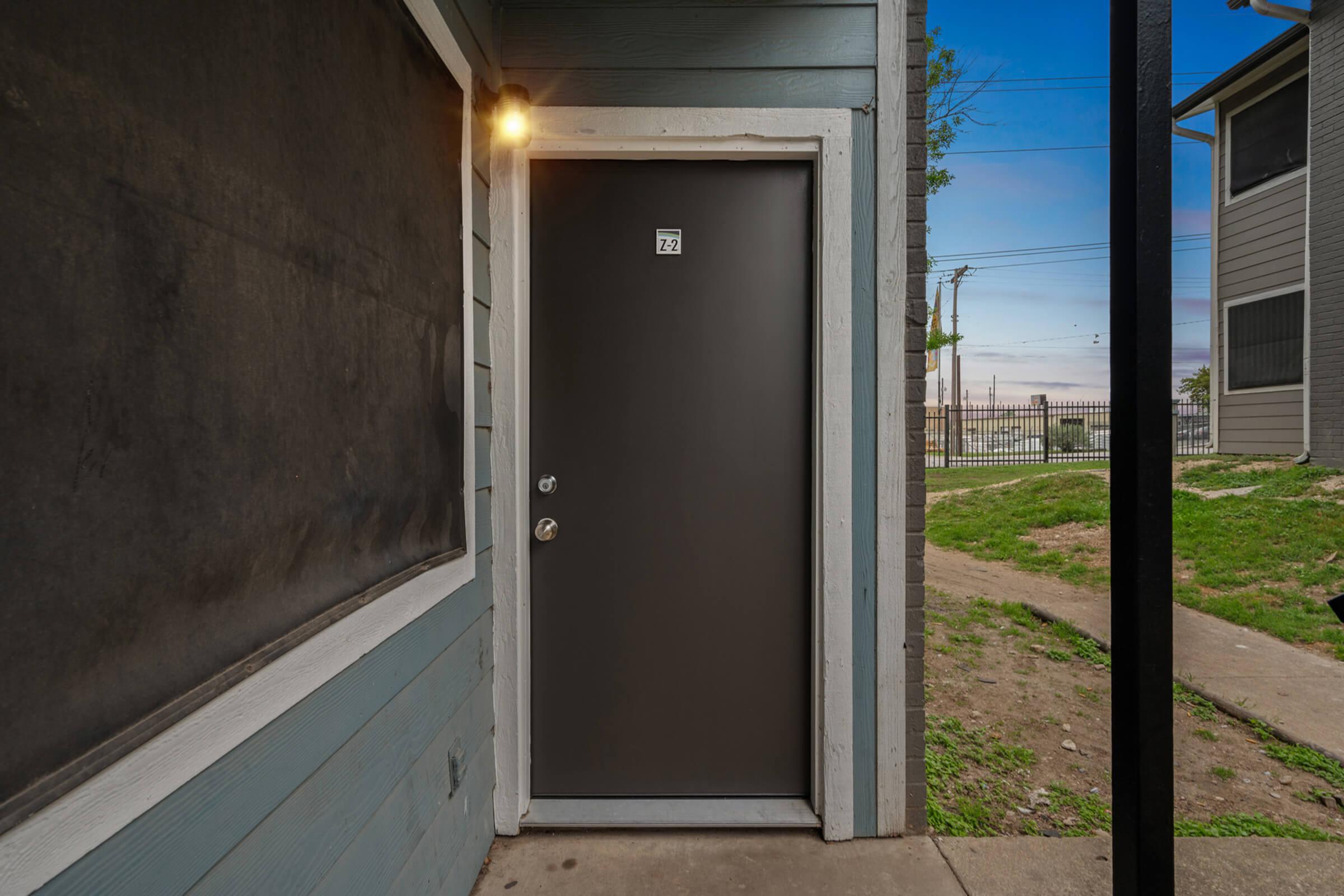 A dark gray entrance door with the number 72 visible is situated on a blue wooden exterior wall. Next to the door, there is a small light fixture. A grassy area and a fence are visible in the background, along with utility poles against a clear sky.