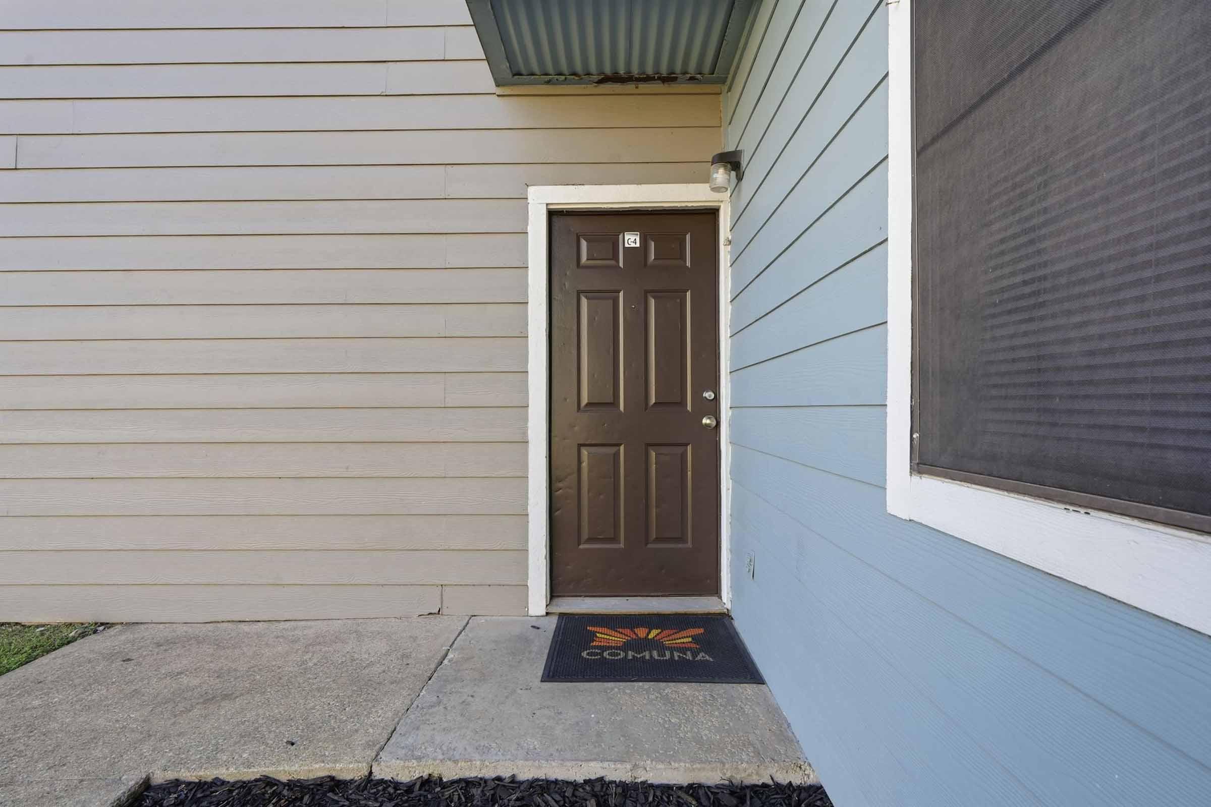 Exterior view of a residential entrance featuring a brown door with a small light above it. The door is flanked by blue siding on one side and a light-colored wall on the other. A welcome mat with the word "COMUNA" is placed at the foot of the door, and the ground is paved with concrete.