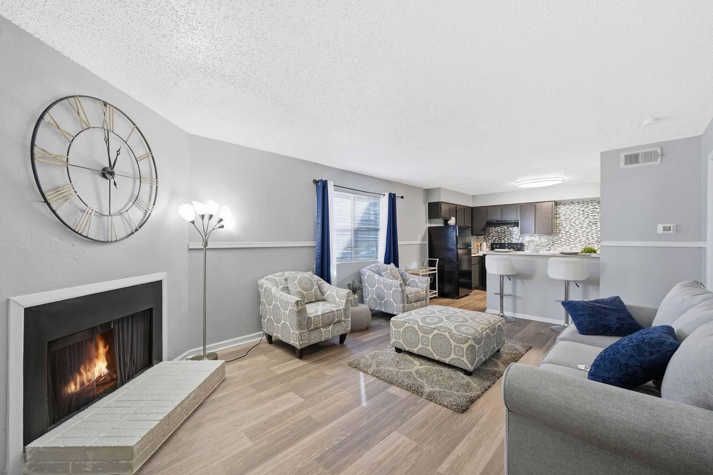 Cozy living room featuring a modern fireplace, two patterned armchairs, and a gray sofa. The room is brightened by natural light through a window with blue curtains. In the background, a kitchen area is visible, complemented by stylish bar stools and a decorative back wall.