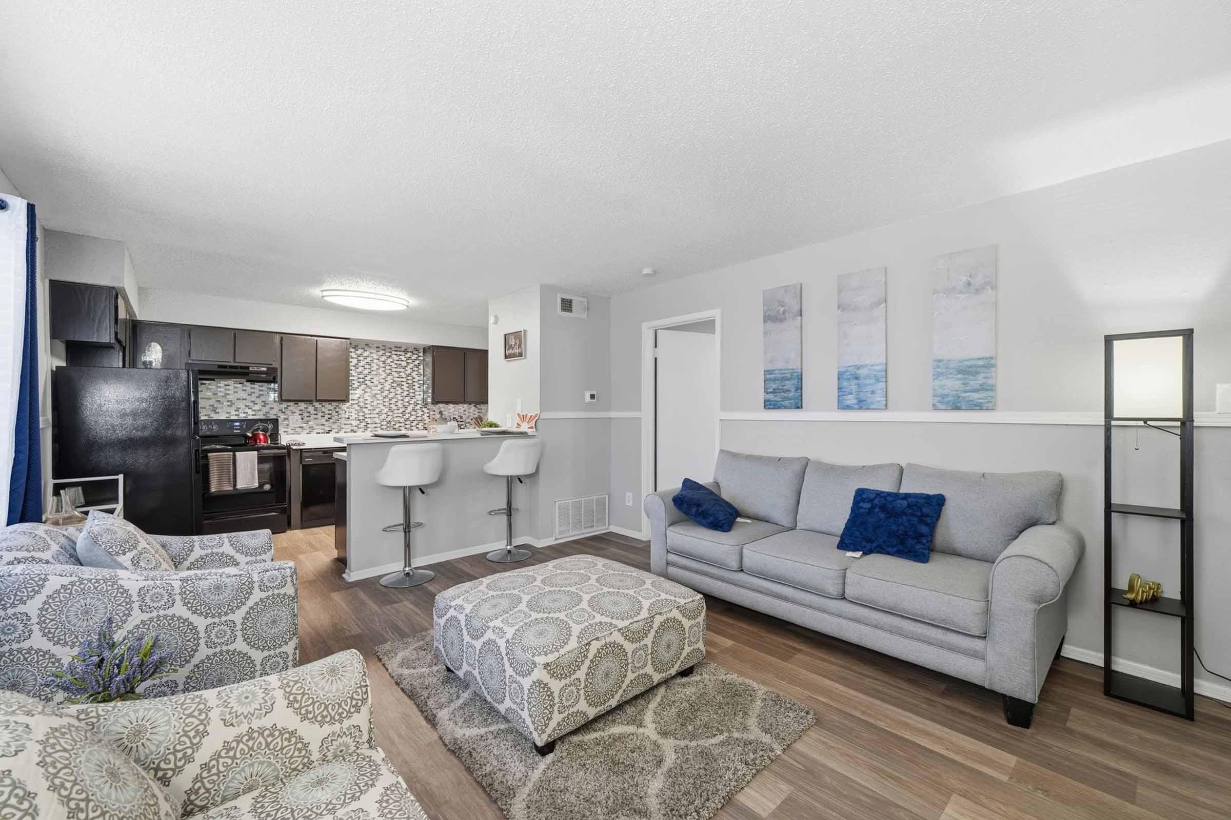A modern living room featuring a gray sofa with pillows, a patterned ottoman, and two patterned chairs. The space includes a dining area with white bar stools and a kitchen in the background with dark cabinetry and light countertops. Natural light enters through windows with blue curtains, enhancing the cozy atmosphere.