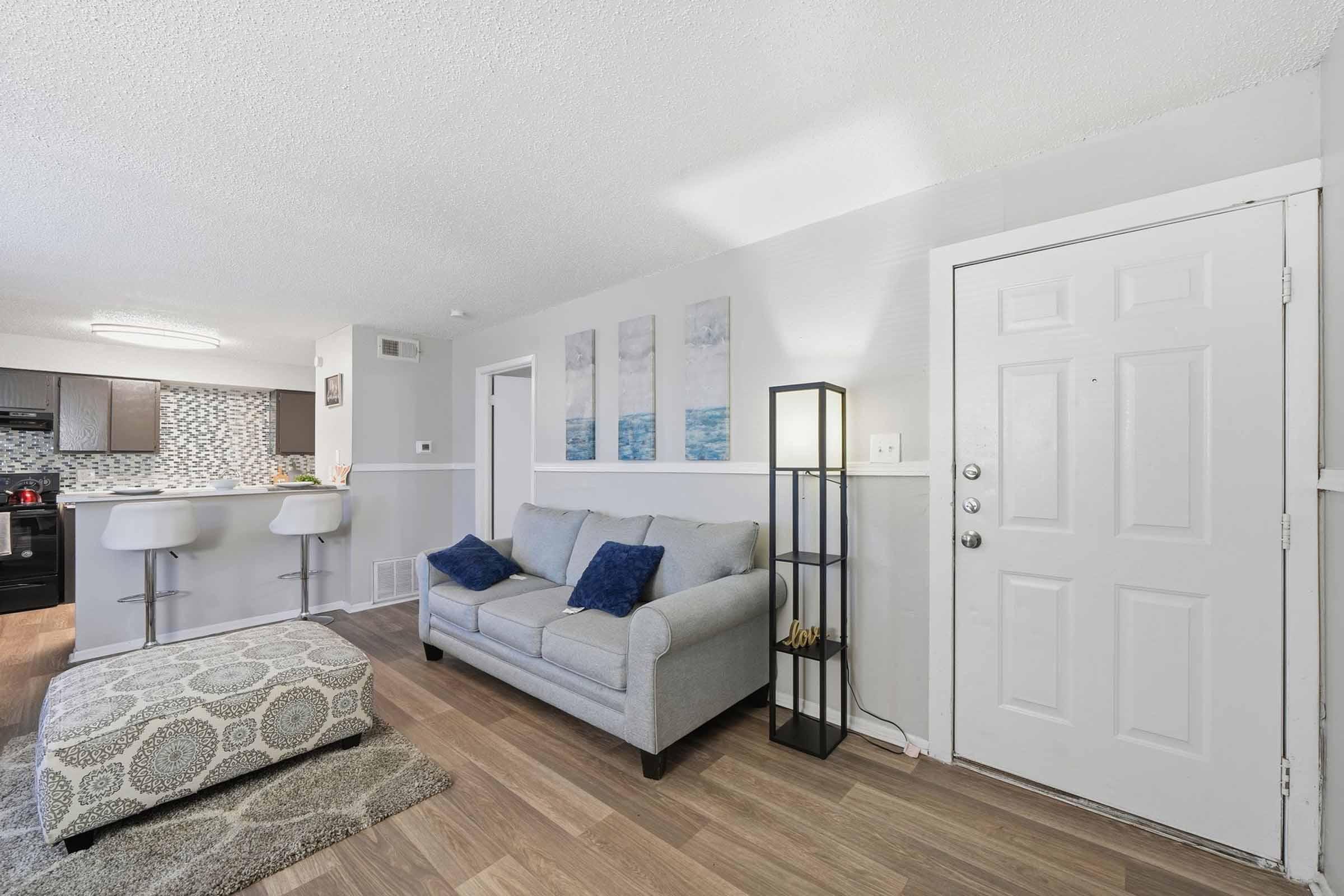 A cozy living room with a light gray sofa adorned with blue pillows, an ottoman with a patterned design, and a standing lamp. The entrance door is visible on the right, and there's a small bar area with white stools in the background, showcasing modern decor with a geometric pattern and artwork on the walls.