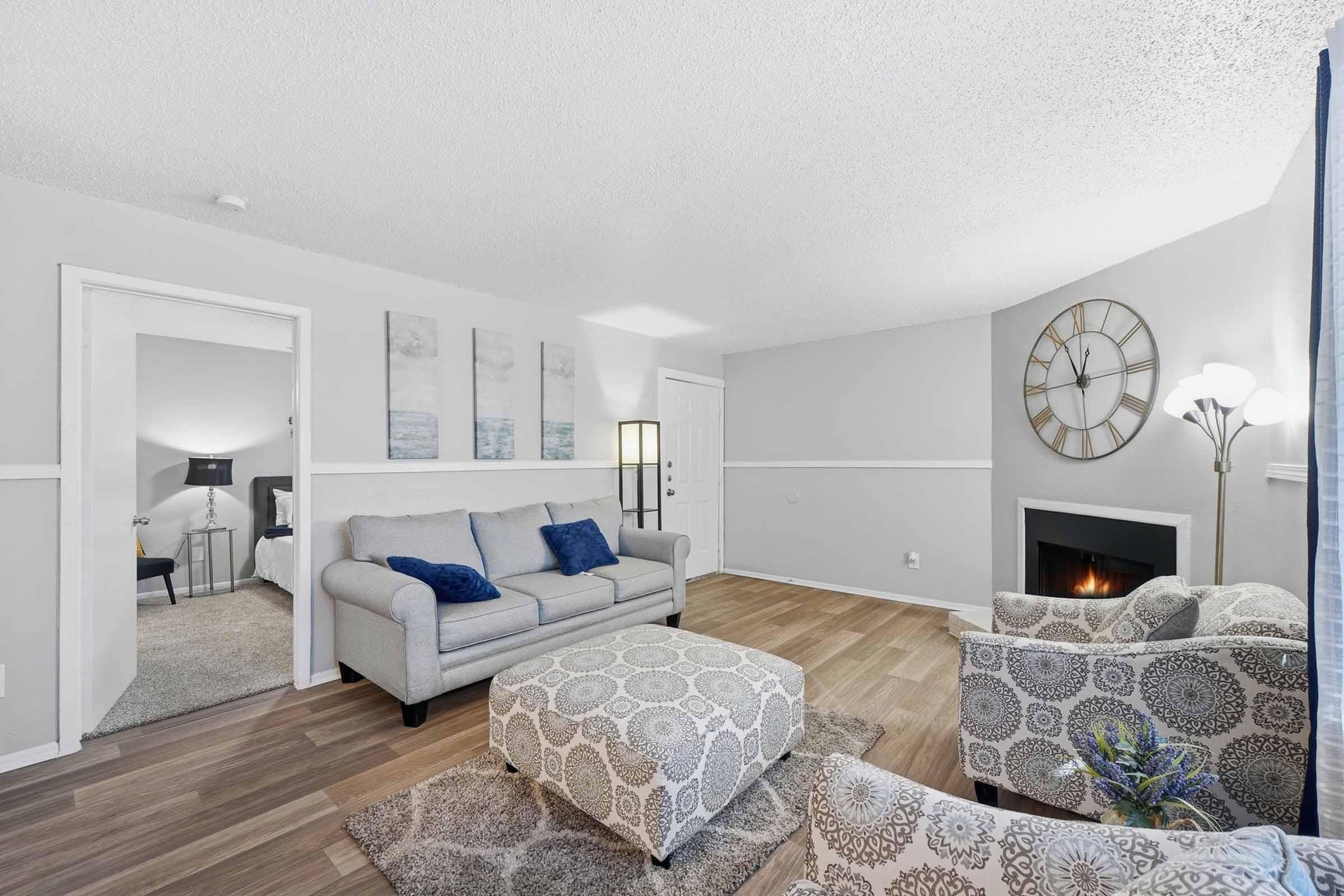 A well-decorated living room featuring a gray sofa with blue cushions, a patterned ottoman, and two cozy armchairs. A stylish lamp stands next to the seating area, and a wall clock adorns one wall. A doorway leads to another room, and a fireplace adds warmth to the space. The walls are painted in neutral tones.