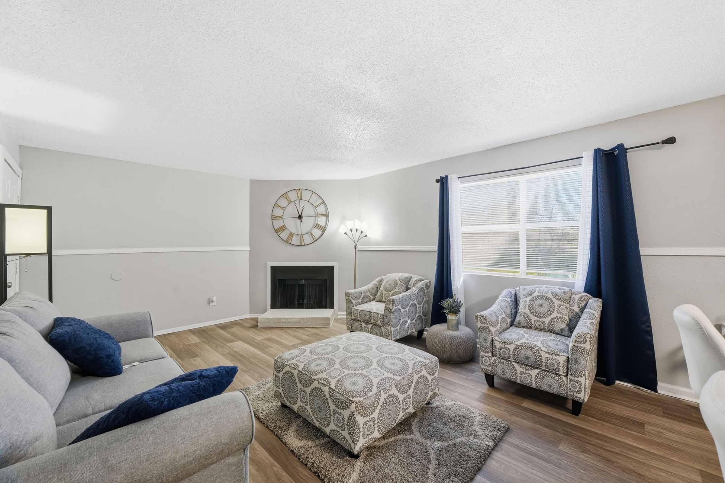A cozy living room featuring a gray sofa with blue and white accent pillows, two patterned armchairs, a decorative ottoman, and a wall clock. The space has large windows with dark curtains, a fireplace, and wood flooring, creating a warm and inviting atmosphere.