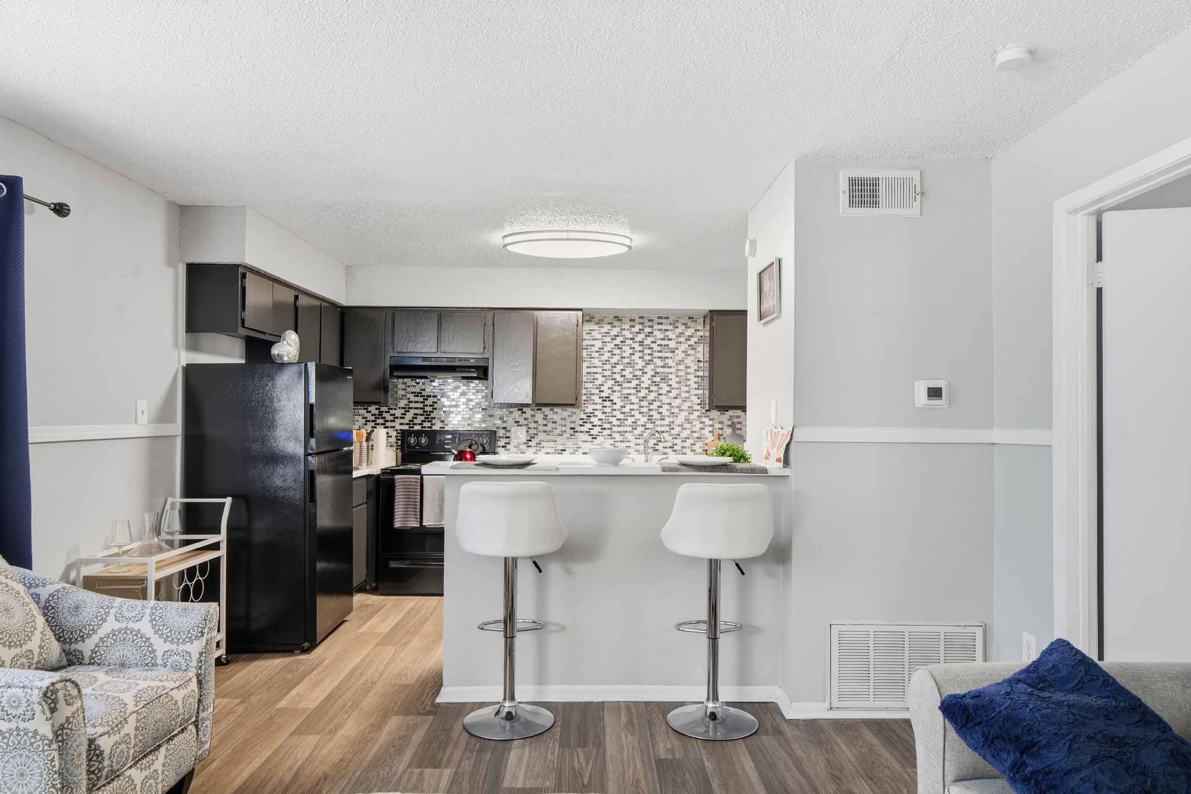 Modern kitchen and living area with a glossy black refrigerator, a light fixture overhead, and a breakfast bar with two white stools. The background features a textured backsplash and dark cabinetry. A cozy seating area is seen with a patterned armchair, enhancing the inviting atmosphere of the space.