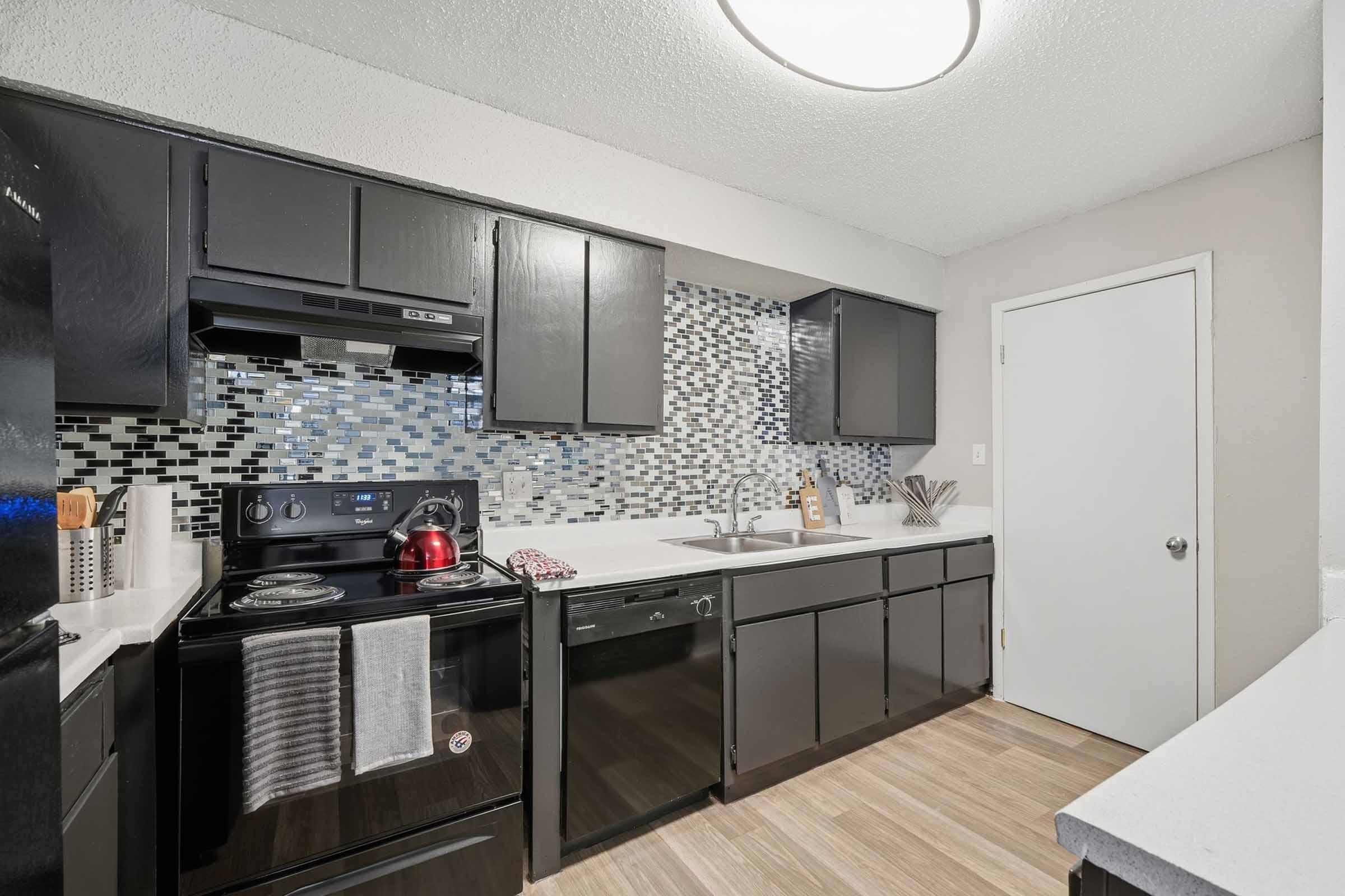 Modern kitchen featuring black cabinets, a glass tile backsplash in shades of gray and blue, stainless steel appliances, and a white countertop. A red kettle sits on the stove, and a door is visible on the right side, adding functionality to the space. Bright overhead lighting enhances the overall ambiance.