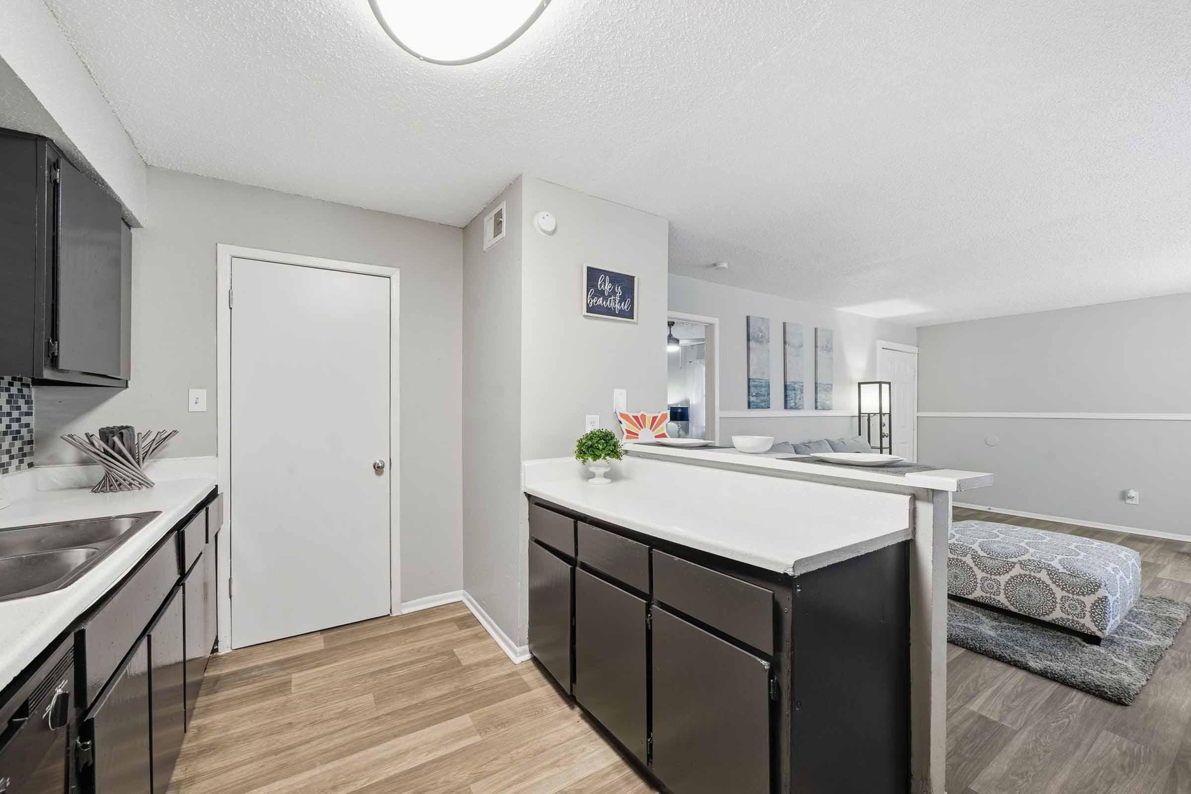 Interior view of a modern apartment featuring a kitchen with black cabinetry and a white countertop, leading to a living area with light-colored walls and decorative artwork. The floor has a wood-like finish, and there's a cozy seating area visible in the background.