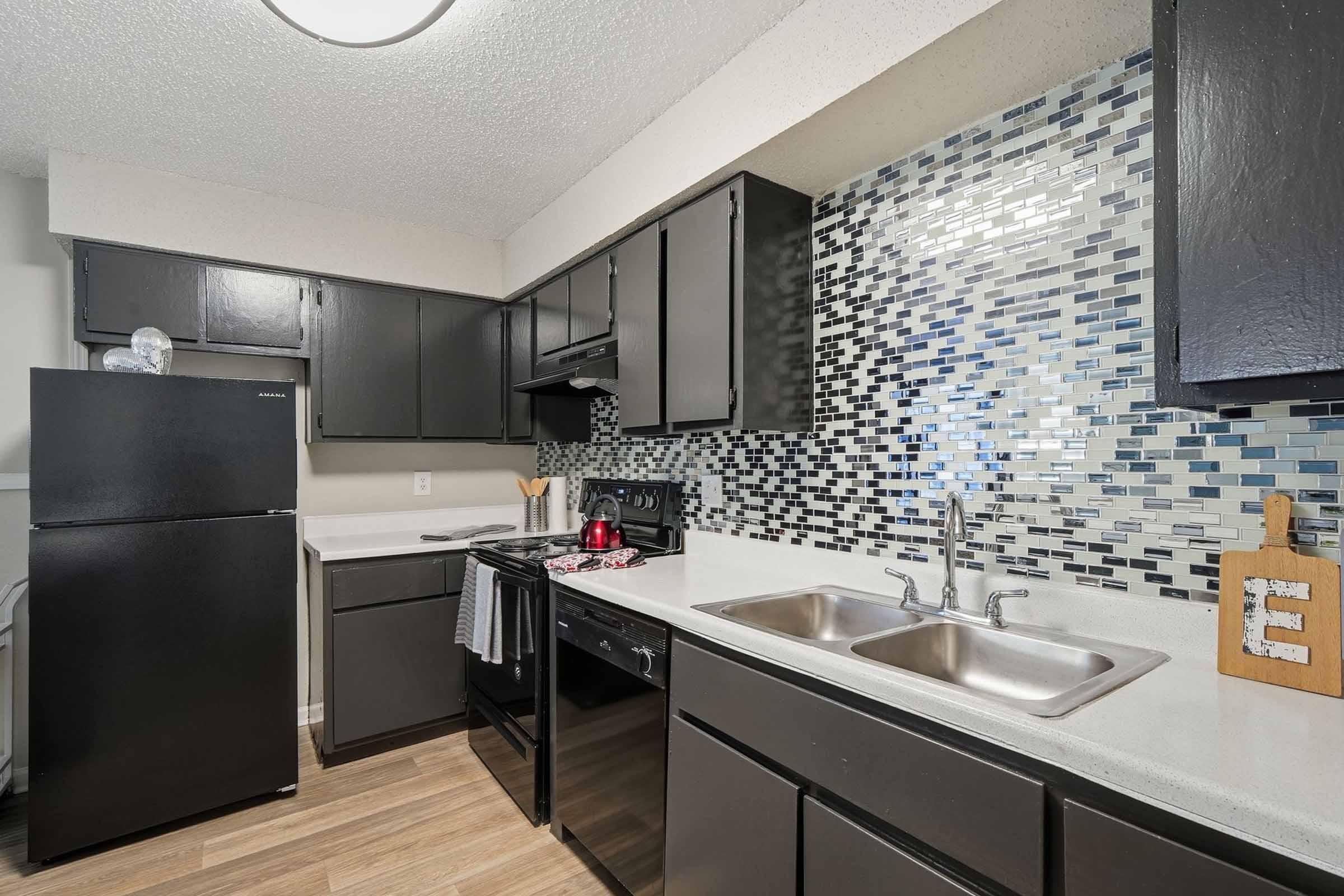 Modern kitchen featuring black cabinets, a black refrigerator, and an electric stove. The backsplash is composed of shiny blue and gray mosaic tiles. A double sink with a chrome faucet is placed under a window, and kitchen utensils are neatly arranged on the countertop. The flooring is light-colored wood.