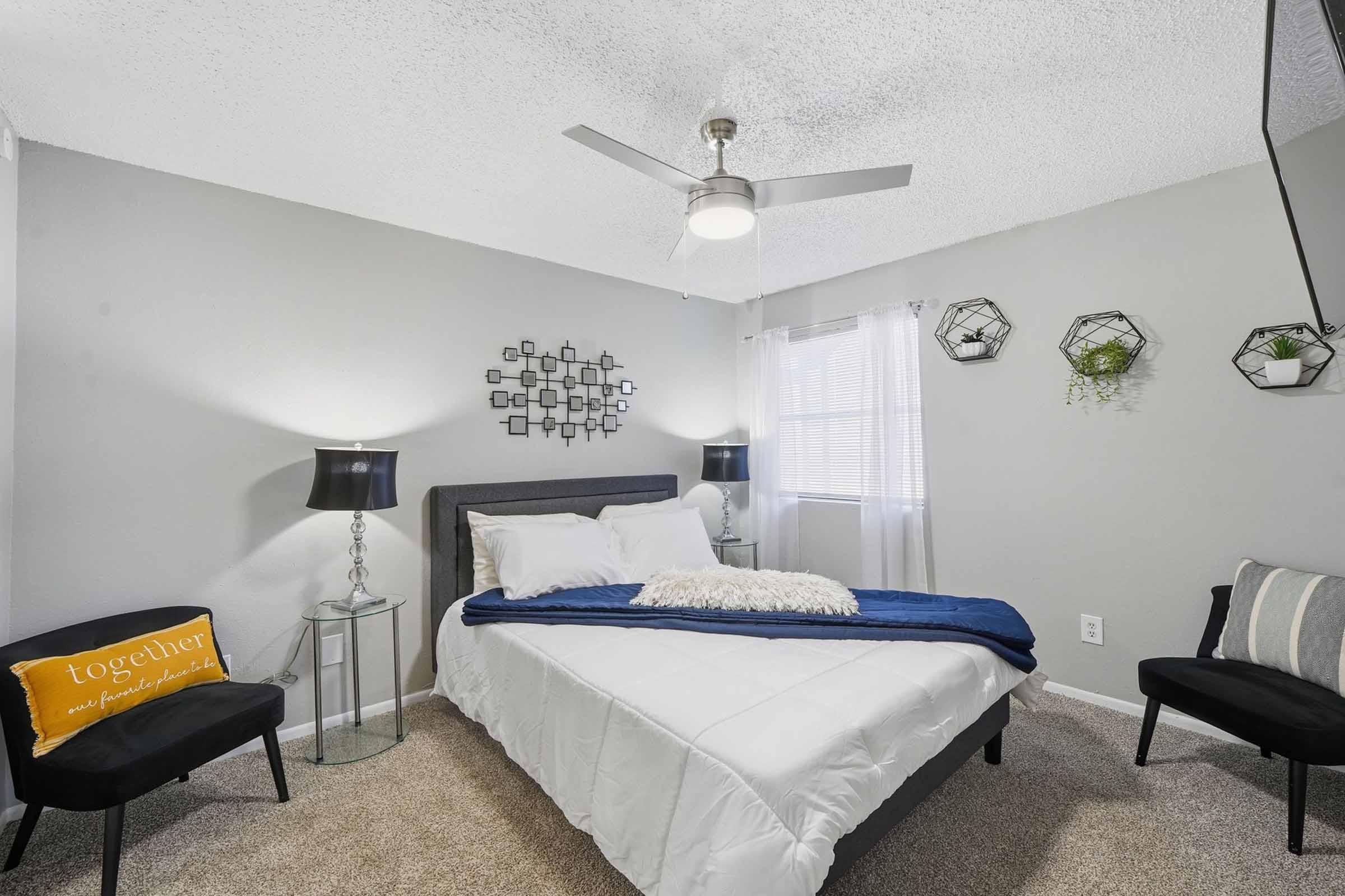 A cozy bedroom featuring a queen-sized bed with a white comforter and decorative blue throw. Two black bedside lamps flank the bed, and a stylish mirror is mounted on the wall. A plant adorns the window, and geometric wall decor adds a modern touch. A black chair and striped cushion complete the inviting atmosphere.