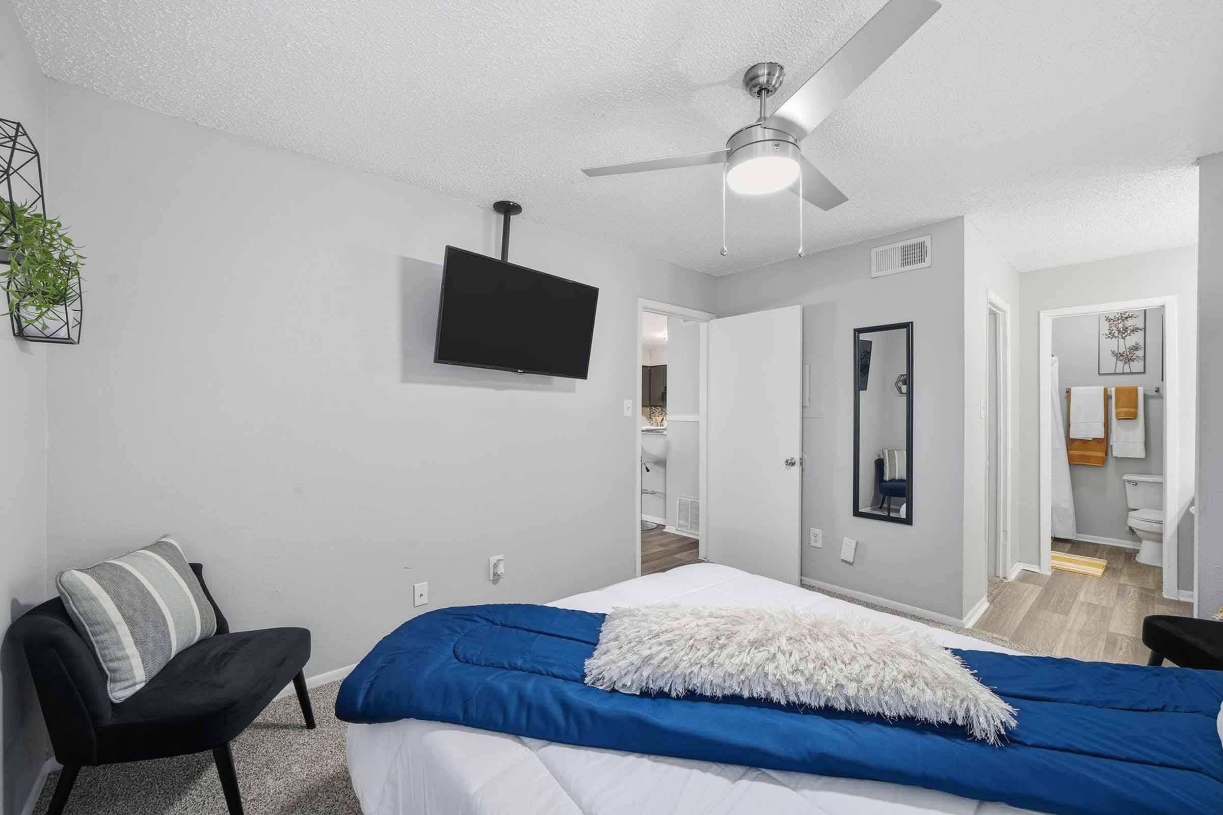 A modern bedroom featuring a queen bed with a blue throw blanket and a fluffy white pillow. There is an armchair in the corner, a ceiling fan, and a wall-mounted TV. A mirror and bathroom door are visible in the background, along with neutral-colored walls and flooring, creating a bright and inviting atmosphere.