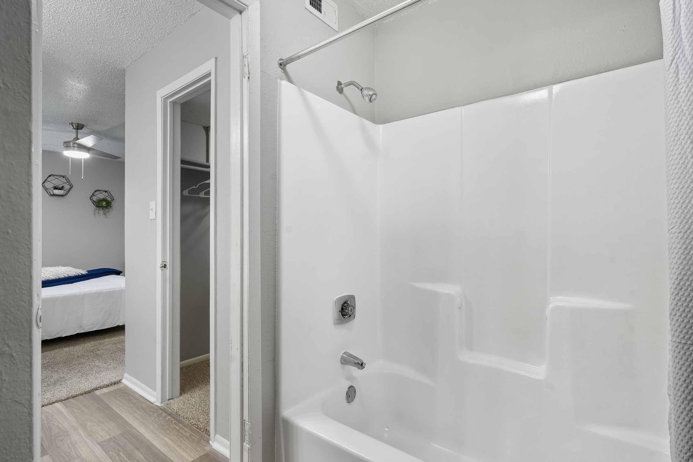 Bathroom view featuring a white shower/tub combination, adjacent to a hallway leading to a bedroom. The bedroom has a light-colored bed and ceiling fan, with a closet visible on the left. Neutral wall colors and flooring create a clean and modern atmosphere.