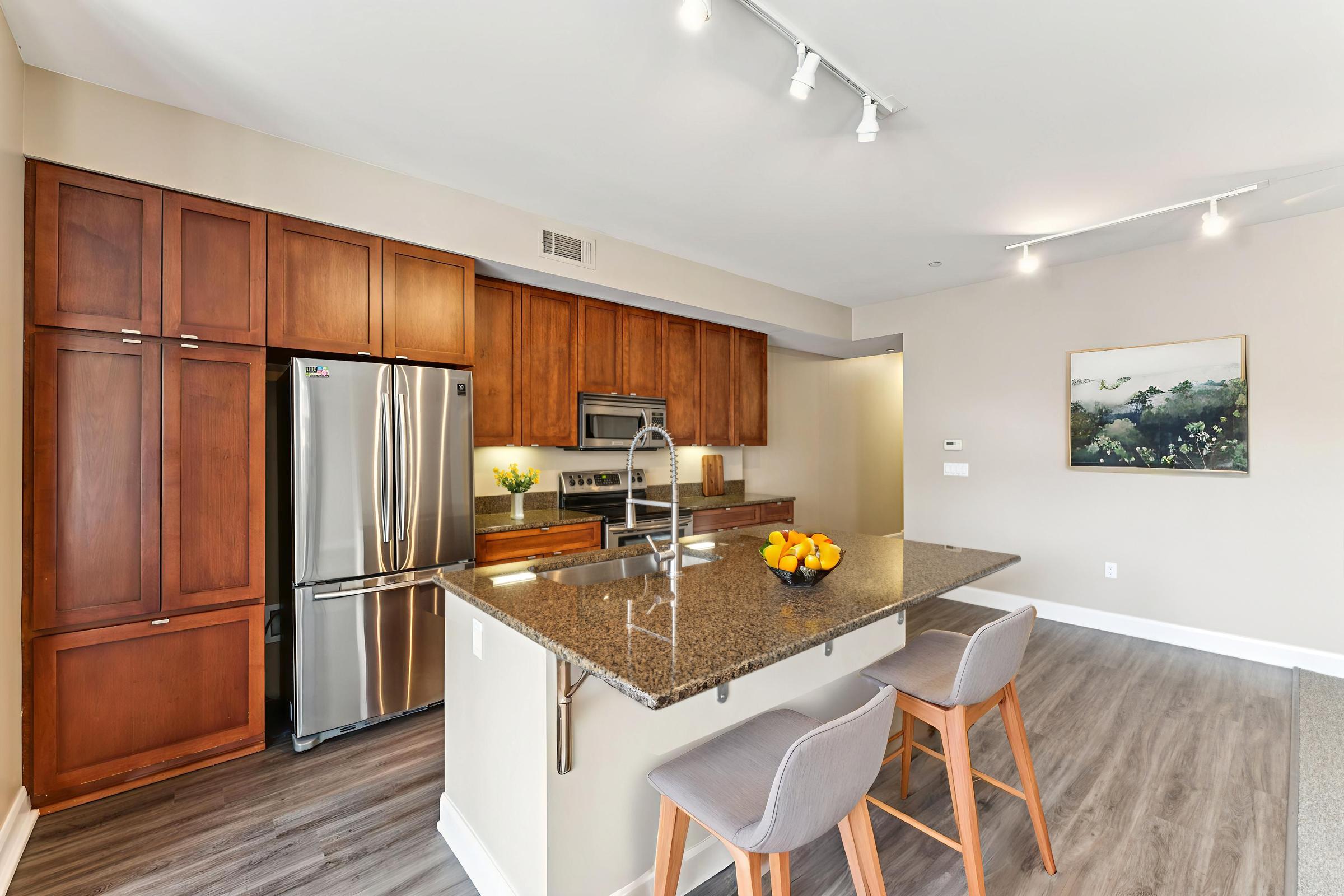Modern kitchen featuring dark wooden cabinets, stainless steel appliances, and a large granite island with seating. The space is well-lit with recessed lighting and has a bowl of oranges on the island. A subtle landscape artwork decorates one wall, complementing the neutral color scheme and contemporary design.