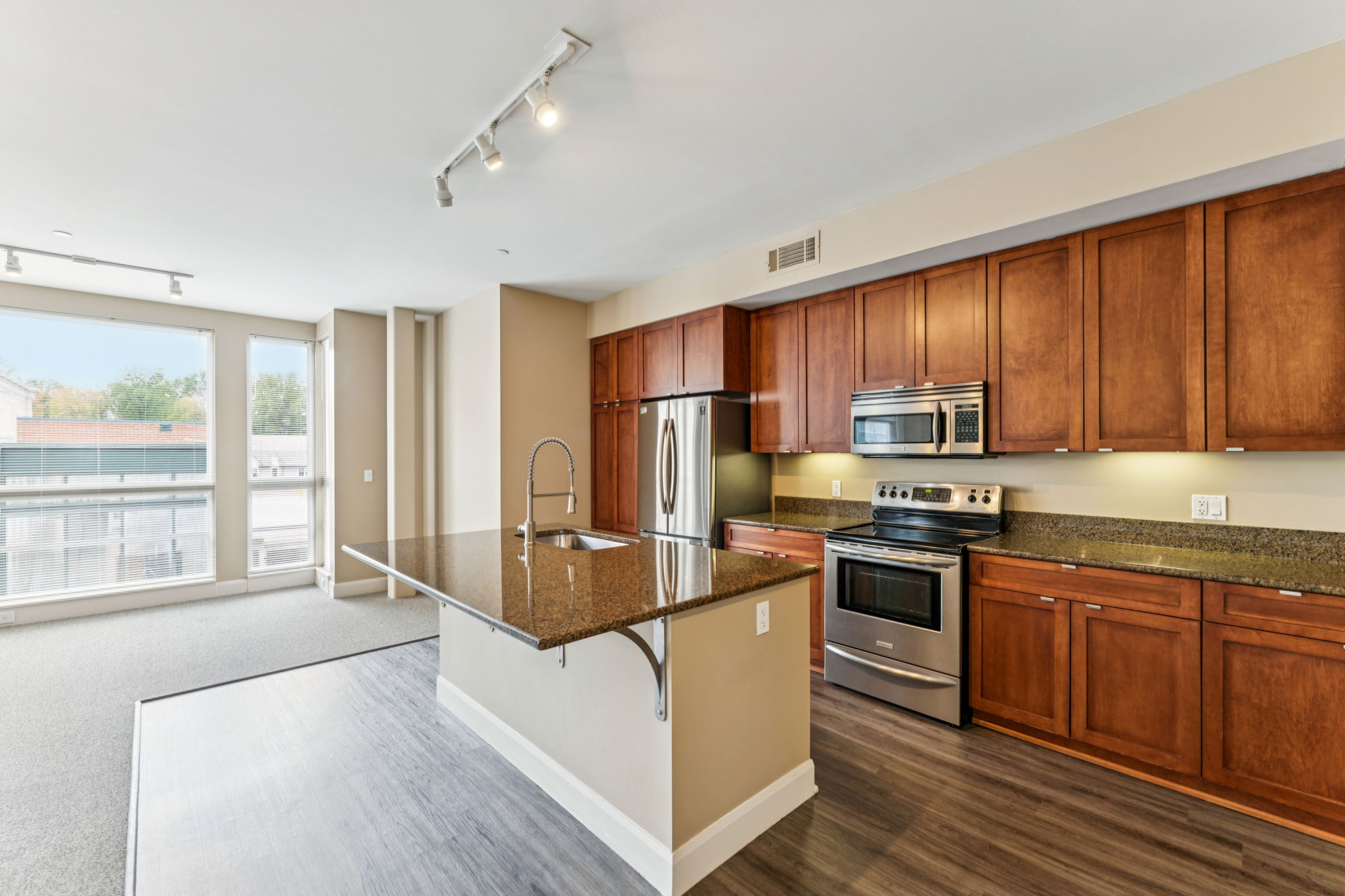 Modern kitchen and living area featuring wooden cabinets, stainless steel appliances, and granite countertops. Large windows provide natural light, with a view of the outdoors. The open layout creates a spacious feel, complemented by neutral-colored walls and flooring.