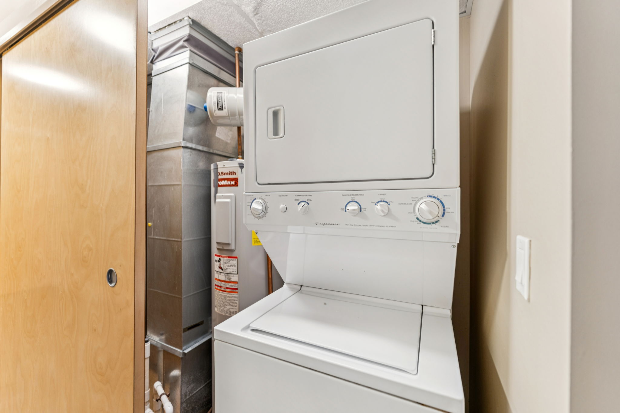 A stacked washer and dryer unit situated in a corner next to a metal vent and a water heater. The appliance features control knobs and a top-loading washer. The surrounding walls are neutral-colored, and a wooden door is partially visible in the background.