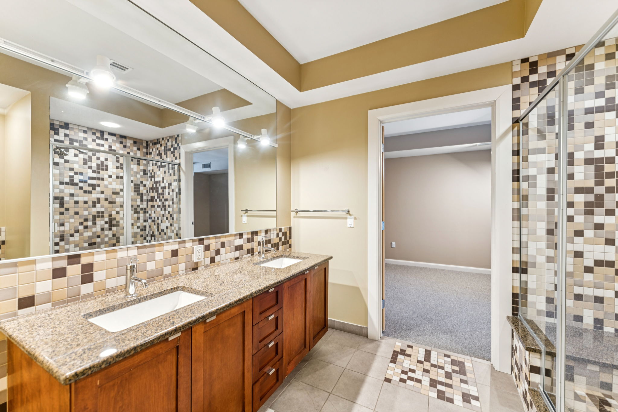 Spacious modern bathroom featuring dual sinks with a granite countertop, large mirror, and contemporary lighting. The walls are adorned with stylish mosaic tiles, and there is a glass-enclosed shower. A doorway leads to a carpeted area outside the bathroom, showcasing a neutral color palette.