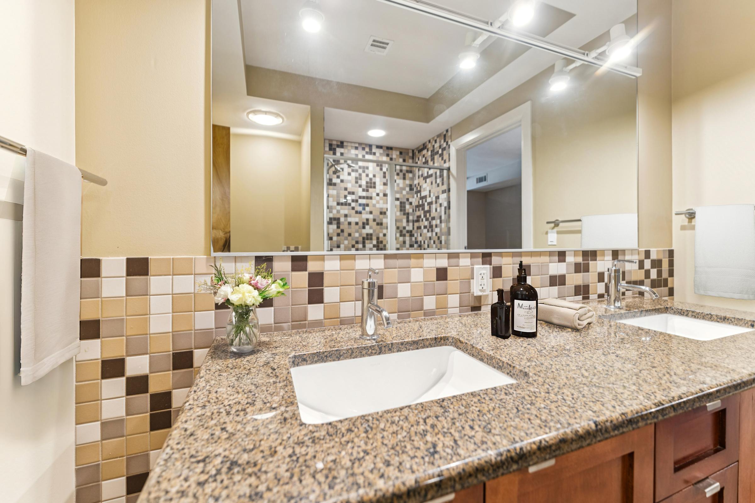 A modern bathroom featuring a granite countertop with a dual sink, a decorative tile backsplash in brown and cream tones, and stylish fixtures. A small vase with flowers sits on the counter, alongside a bottle and a rolled towel, while a glass shower with mosaic tiles is visible in the background.