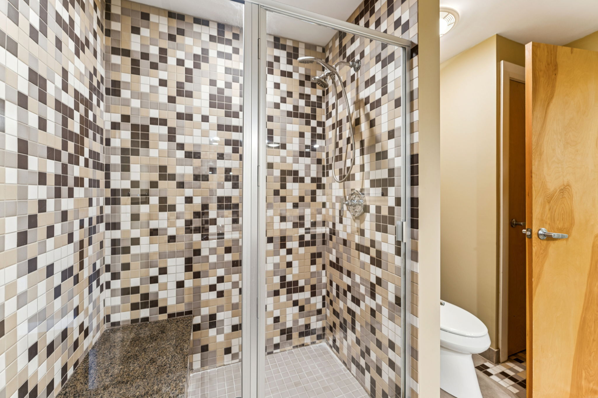 A modern shower stall featuring a glass enclosure, surrounded by a vibrant mosaic tile pattern in shades of brown, cream, and gray. A sleek showerhead is mounted on the wall, and the space includes a small ledge with a dark surface. A door to the right leads to another area.