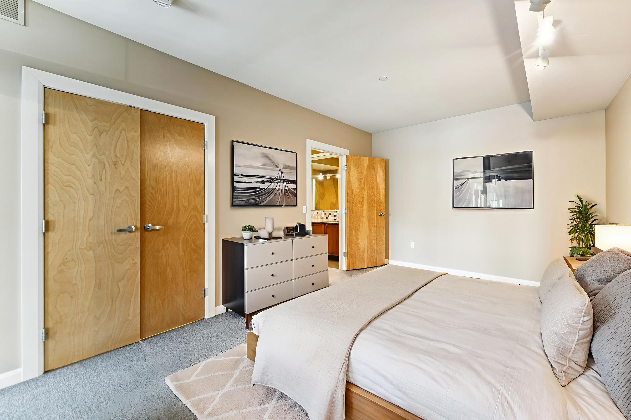 A modern bedroom featuring a double bed with neutral bedding, a dresser, and two wooden doors leading to a bathroom. Decorative artwork adorns the walls, and a small plant adds a touch of greenery. The room has soft lighting and a cozy atmosphere, with light-colored walls and carpet.