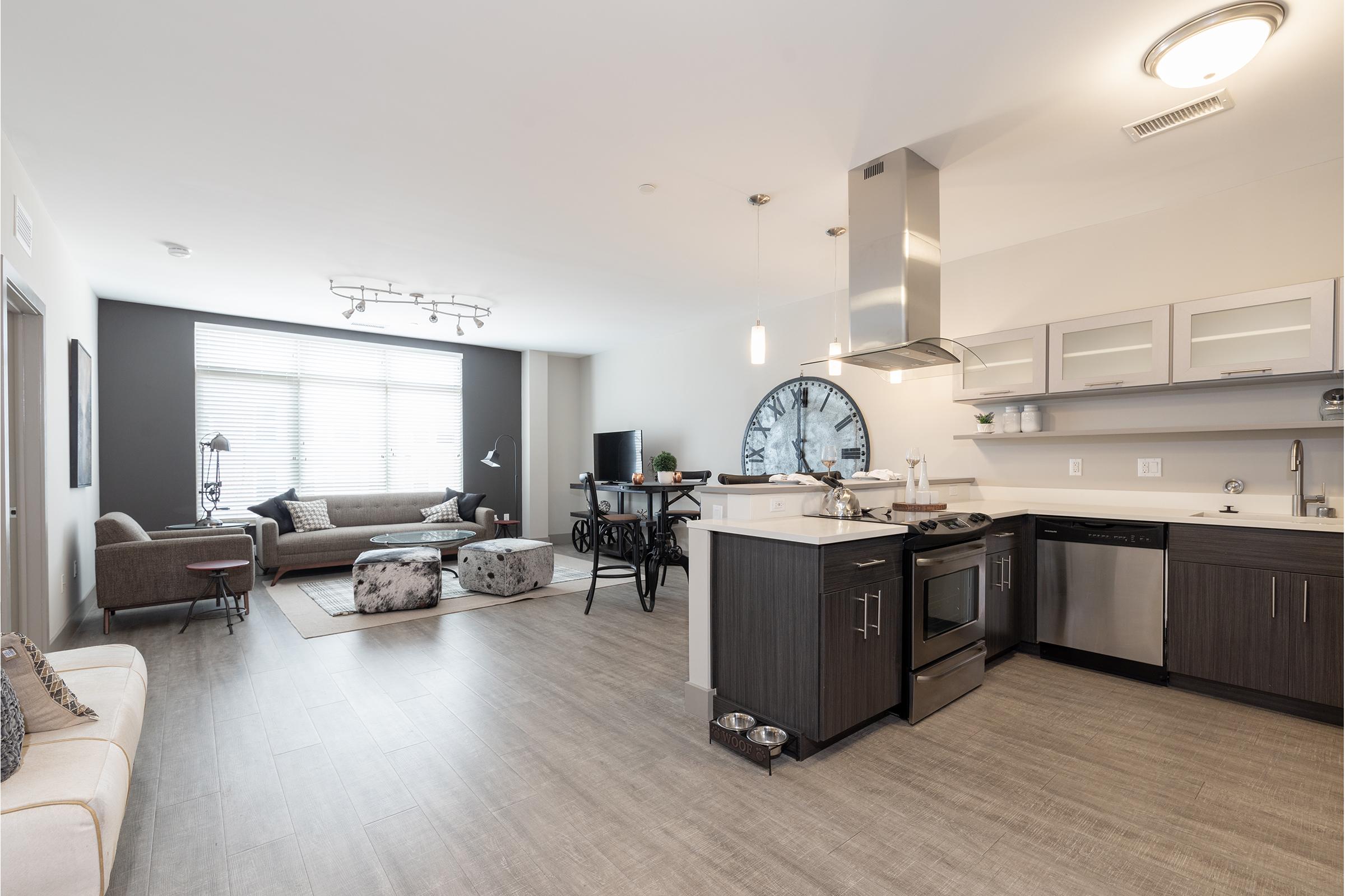 Modern open-concept living space featuring a kitchen with stainless steel appliances, a dining area with a large clock, and a cozy living room with comfortable seating and natural light from large windows. The decor includes neutral colors and stylish furnishings.