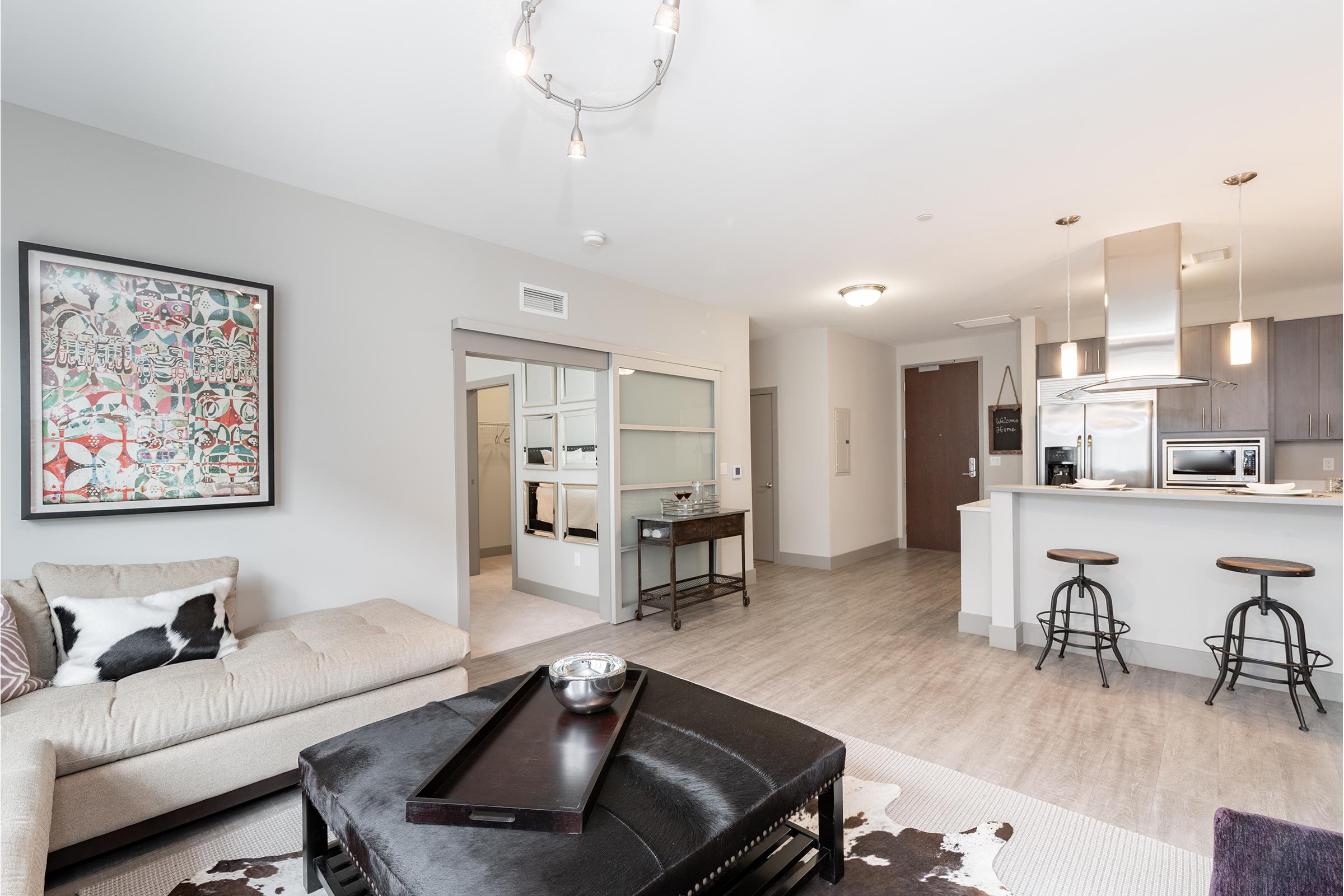 A modern living space featuring a cozy beige sofa with a decorative black and white pillow, a stylish coffee table, and a sleek kitchen visible in the background. The decor includes artwork on the wall, a light fixture above, and an open layout with wooden flooring.