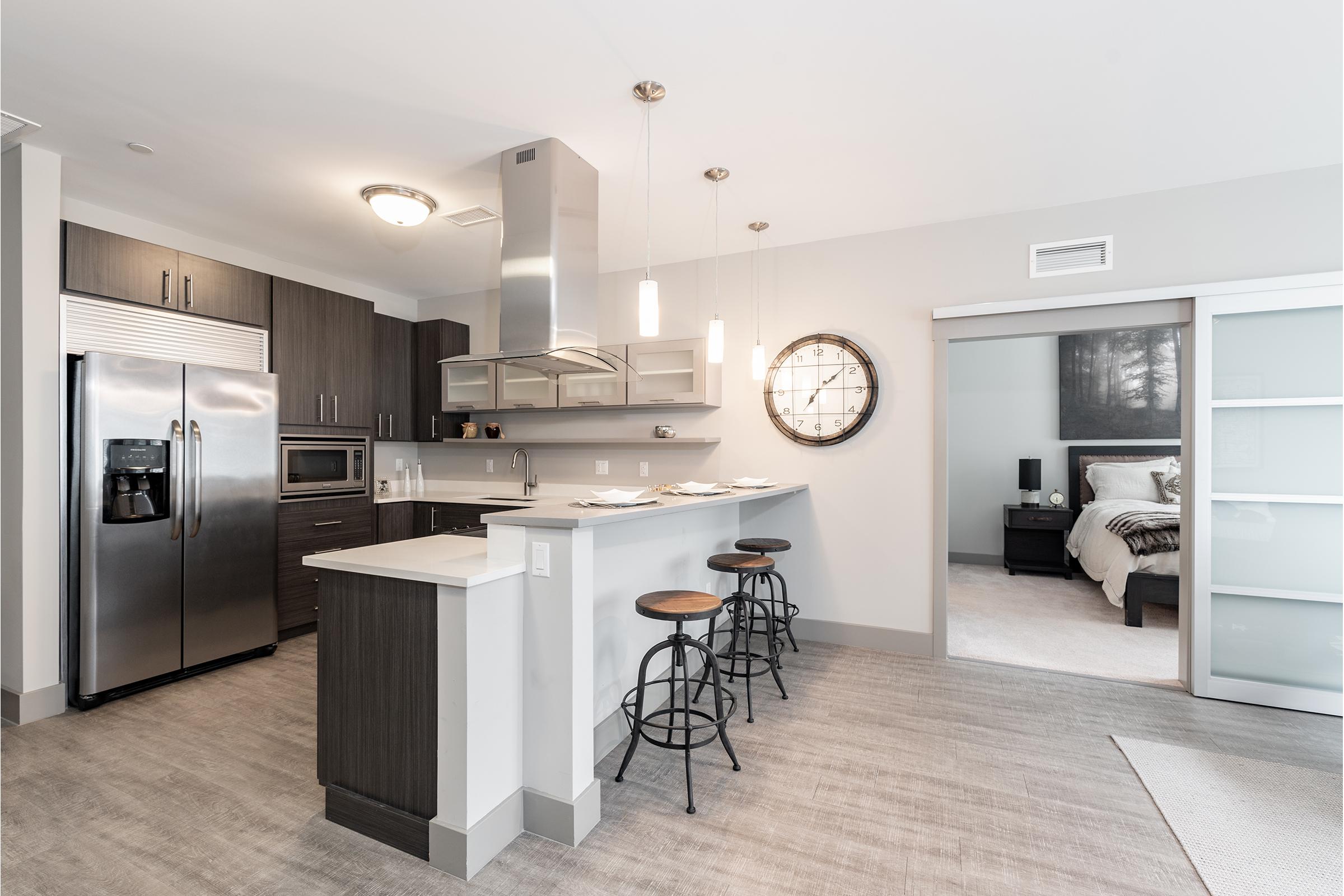 Modern kitchen with stainless steel appliances, including a large refrigerator and oven. Features a sleek island with three black bar stools, grey cabinetry, and a unique wall clock. A sliding glass door leads to a bedroom area with light-colored decor. The space is bright and contemporary.