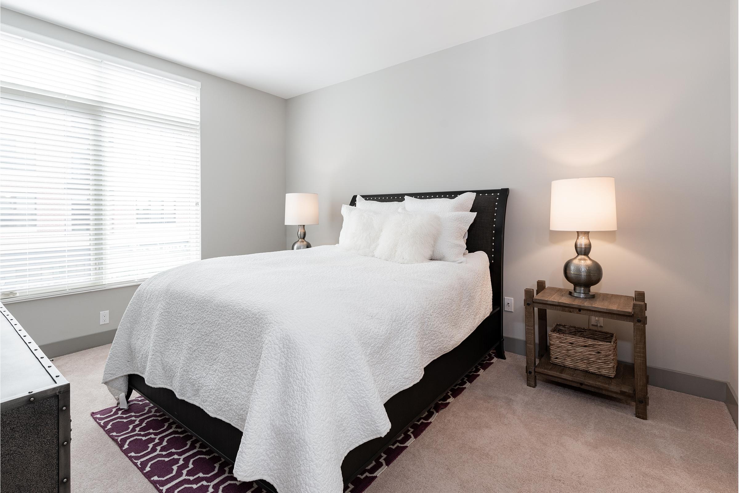 A cozy bedroom featuring a queen-sized bed with white bedding and plush pillows. Flanking the bed are two stylish bedside lamps on wooden tables. A soft area rug in purple adds color to the light-toned carpet. Natural light streams through a window with horizontal blinds, creating a warm and inviting atmosphere.