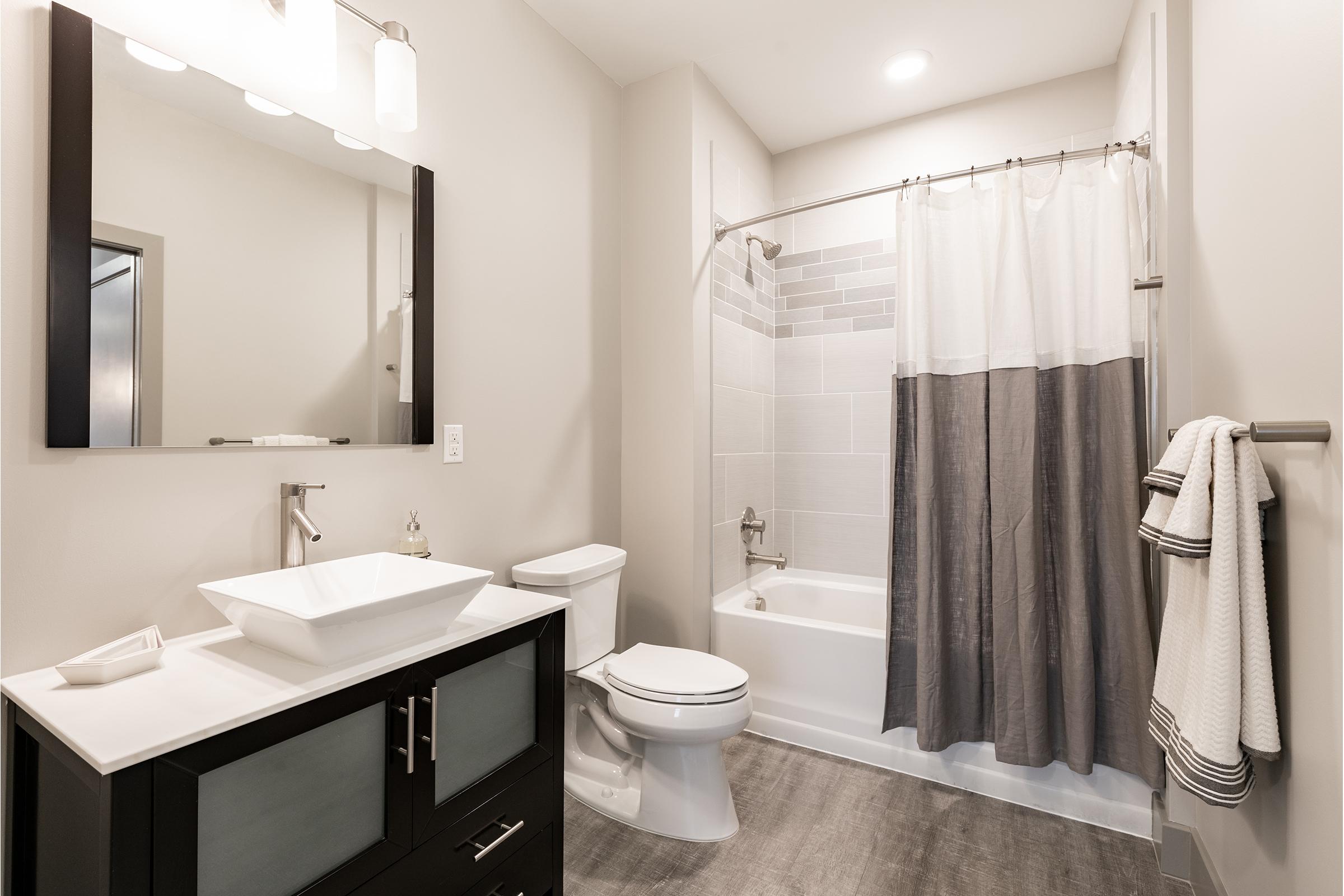 Modern bathroom featuring a sleek design with a black vanity, white sink, and a large mirror. The shower area includes a gray and white curtain, and the space is well-lit with a ceiling light. Neutral walls and a light gray floor complete the contemporary aesthetic. A towel hangs on a nearby rack.