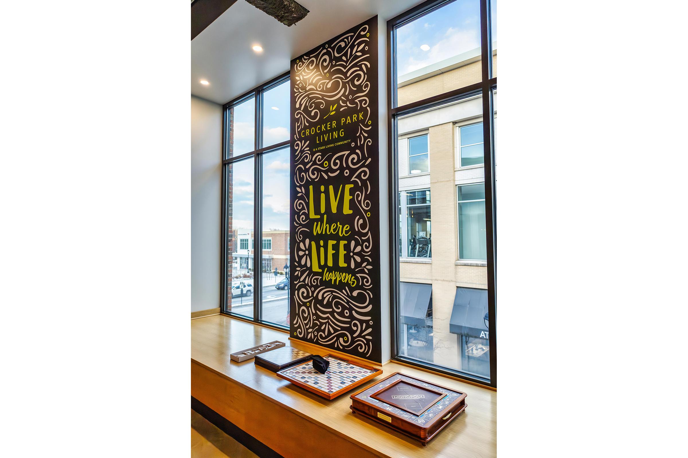 A large wall mural at Crocker Park Living featuring decorative text that reads "Live where life happens." The mural is surrounded by large windows, allowing natural light to fill the space. Below the mural, there are wooden tables with games, creating an inviting atmosphere.