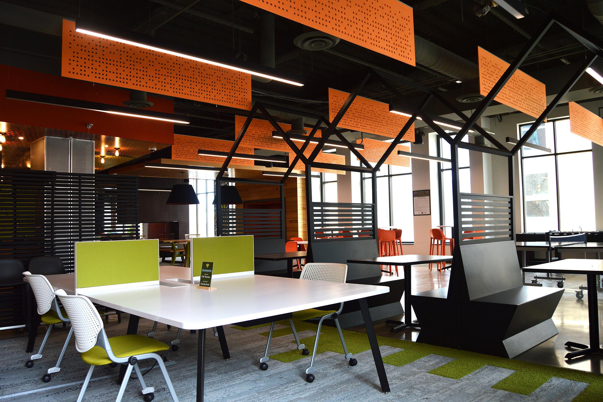 Modern office interior featuring a spacious layout with white tables, colorful chairs in orange and green, and decorative orange panels hanging from the ceiling. The design includes black dividers and a well-lit environment, promoting a collaborative and creative workspace.