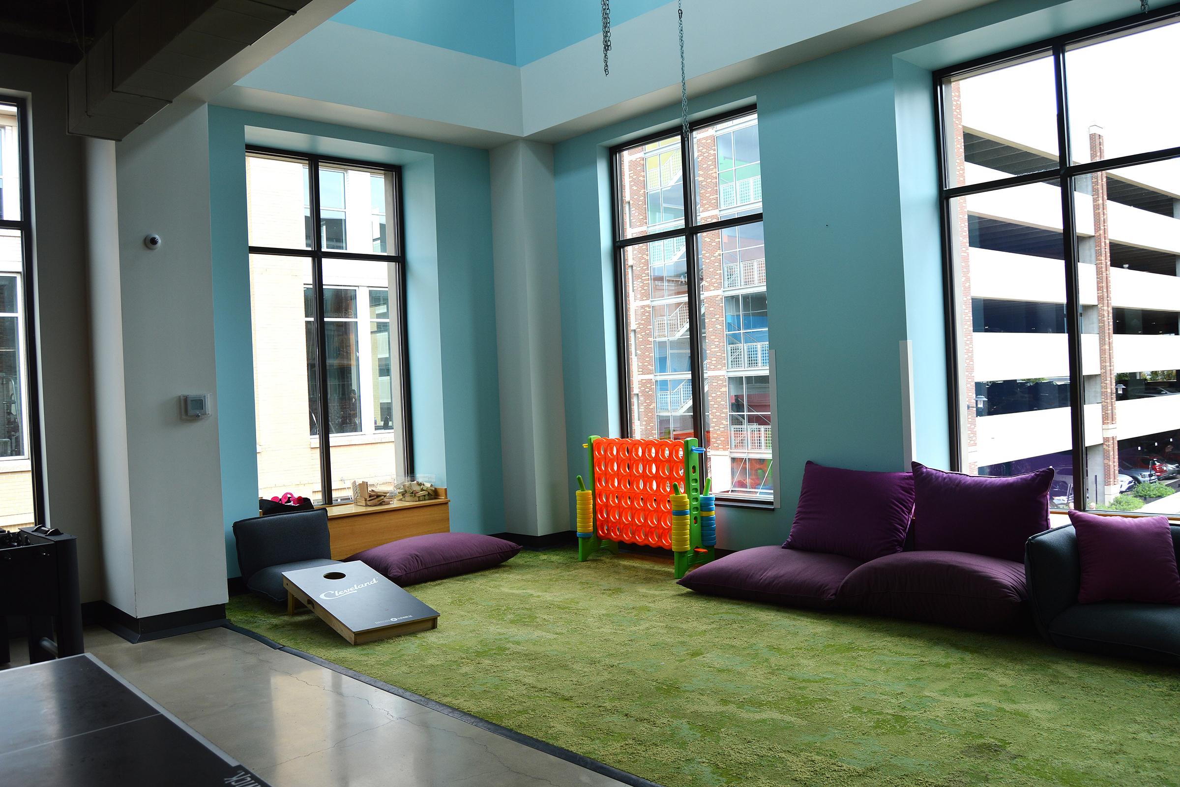 A bright indoor space featuring large windows with a view of a parking lot. The floor is covered in green carpet, and there are purple cushions and a colorful play structure in the corner. A cornhole game board is placed nearby, creating a relaxed and inviting atmosphere for leisure activities.