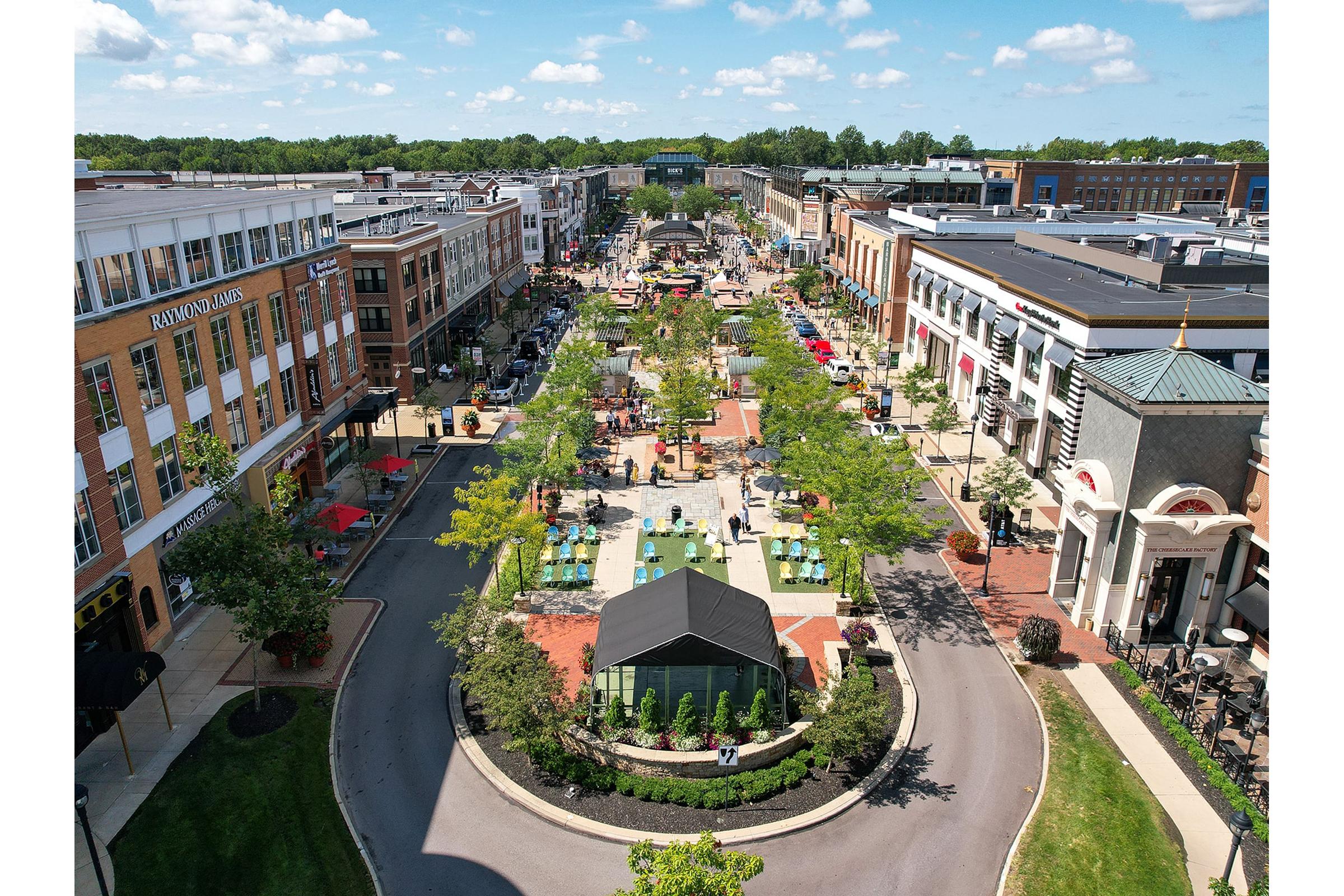 Aerial view of a vibrant outdoor shopping plaza with a wide pedestrian walkway. Lush greenery, trees, and colorful planters line the street. Shoppers and diners enjoy the sunny day, with various storefronts visible. The scene captures a lively urban atmosphere with buildings and outdoor seating areas.