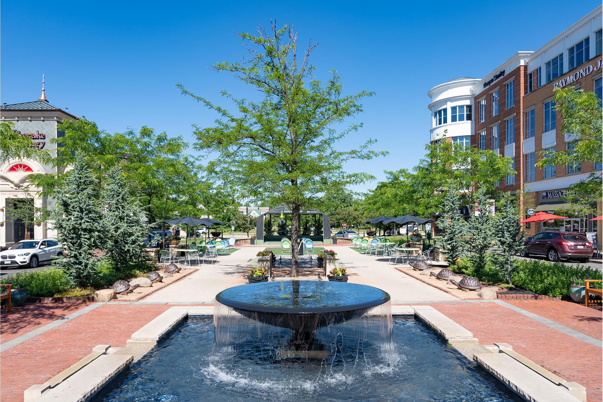 A modern outdoor plaza featuring a central fountain surrounded by greenery and landscaped areas. The scene includes outdoor seating with umbrellas, decorative trees, and nearby buildings in a bright blue sky. The atmosphere is vibrant and welcoming, perfect for leisure and social gatherings.