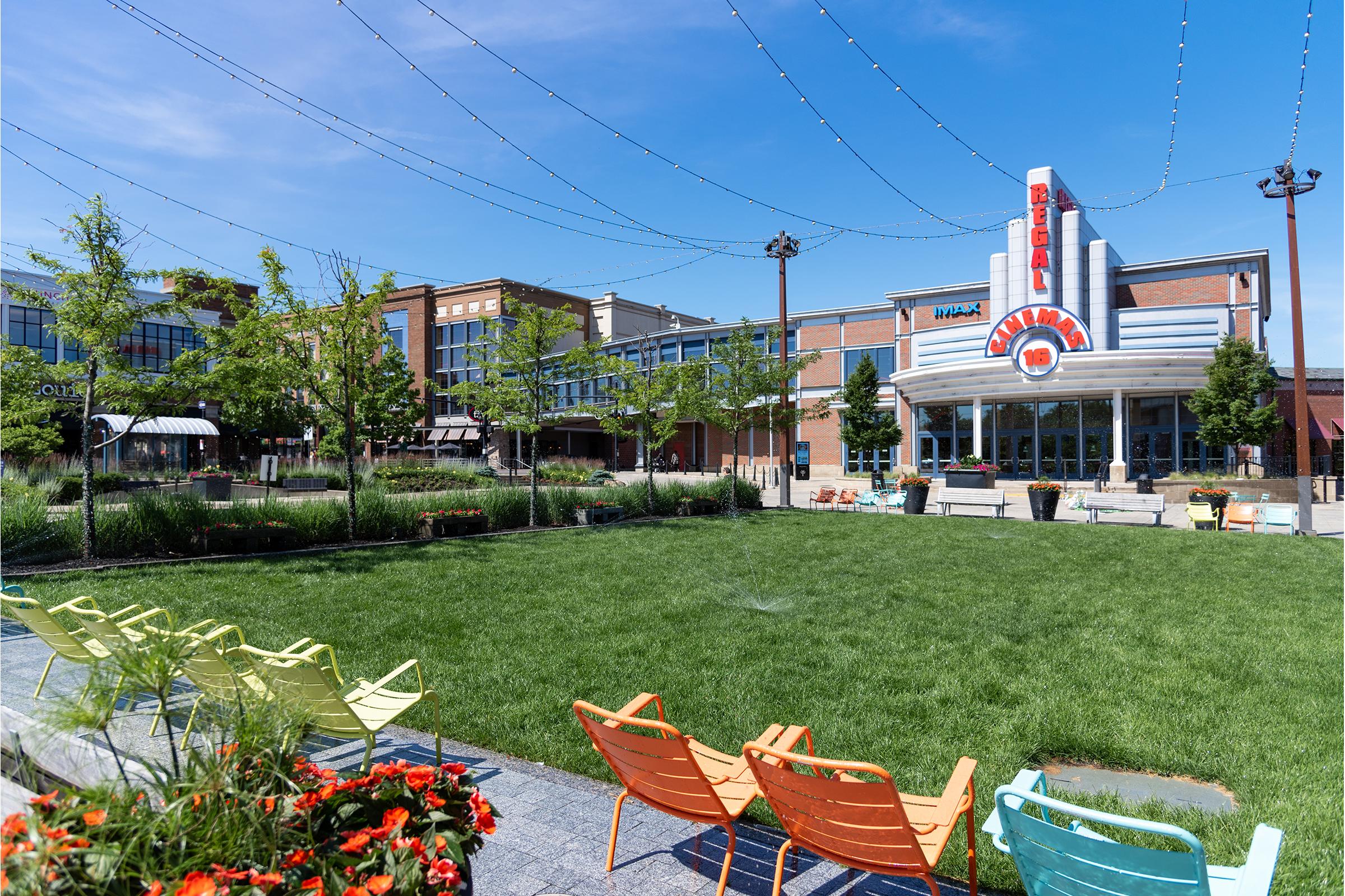 A vibrant outdoor plaza with green grass and colorful seating in the foreground. In the background, a building featuring large signage and modern architecture, accompanied by trees and string lights overhead, creating a welcoming atmosphere. Clear blue sky enhances the lively setting.