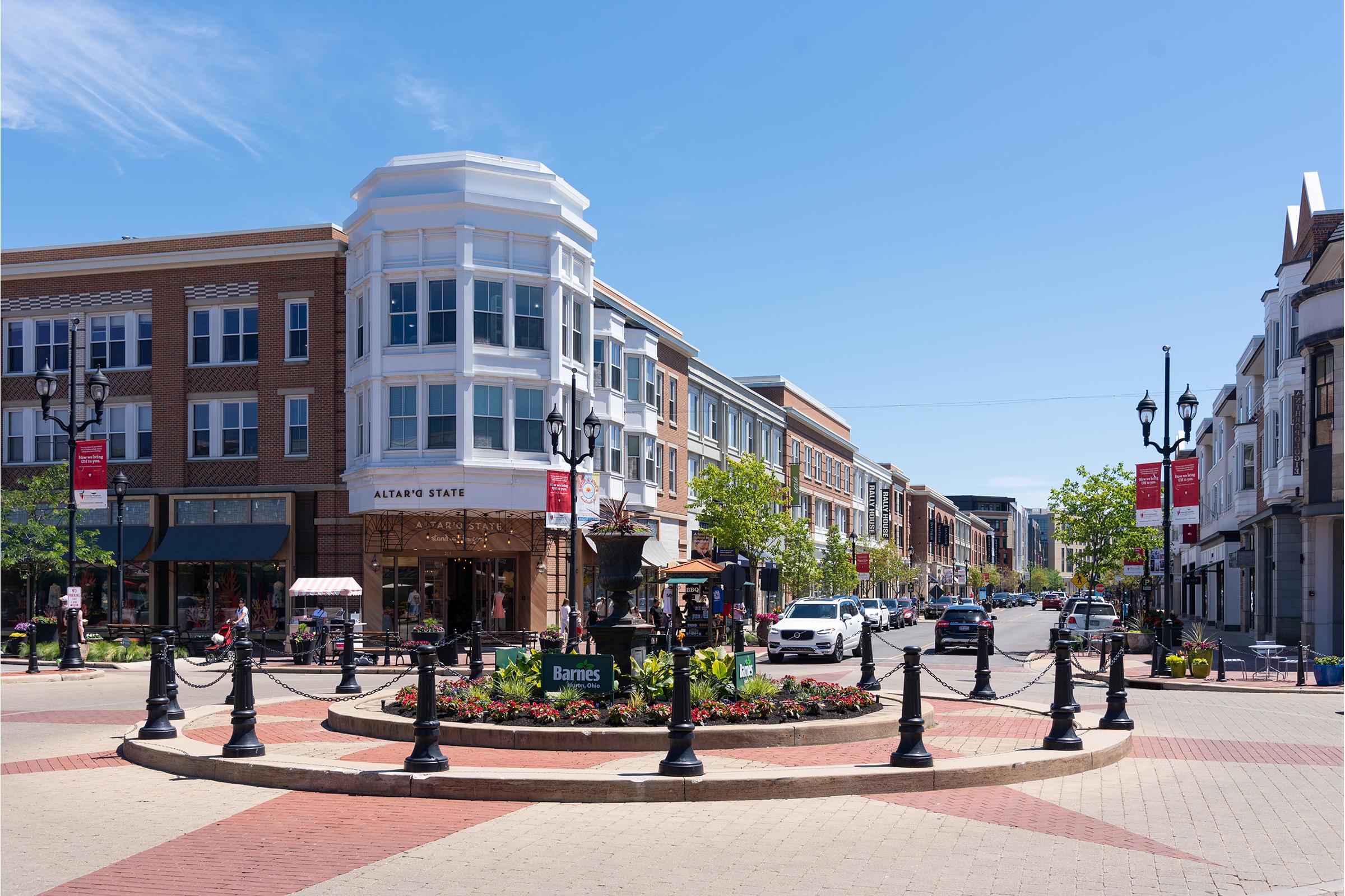 A vibrant urban plaza featuring a circular flower bed at the center, surrounded by decorative fencing. Modern storefronts line the streets, with a bright blue sky overhead. Cars are parked along the road, and people can be seen walking nearby, enjoying the lively atmosphere.