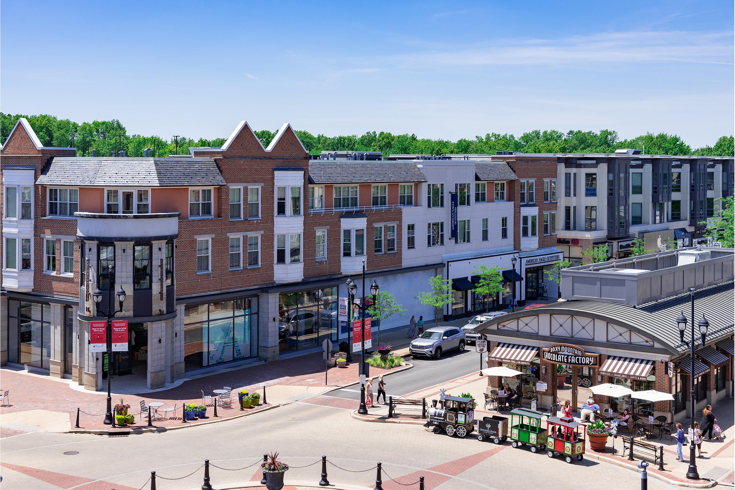 A vibrant outdoor shopping area featuring a mix of brick and modern buildings, with shops and eateries. The street is lined with trees and includes a small market or food stall. Bright, sunny day with clear blue skies enhancing the lively atmosphere.