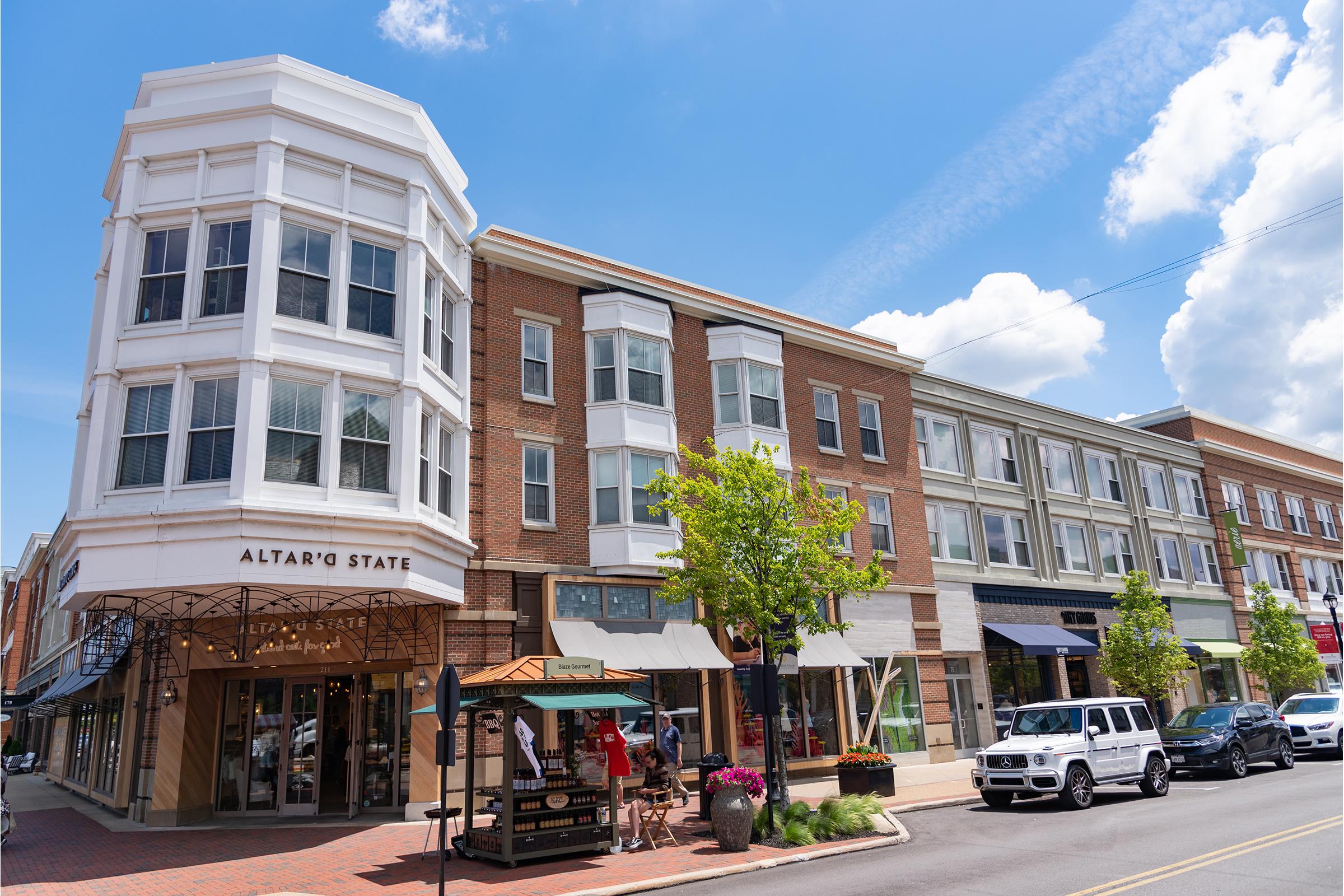 A vibrant street scene featuring modern architecture with a mix of brick and glass facades. The building on the left houses "Altar'd State," while other shops line the street. Planters with colorful flowers and parked vehicles enhance the inviting atmosphere under a bright blue sky with fluffy white clouds.