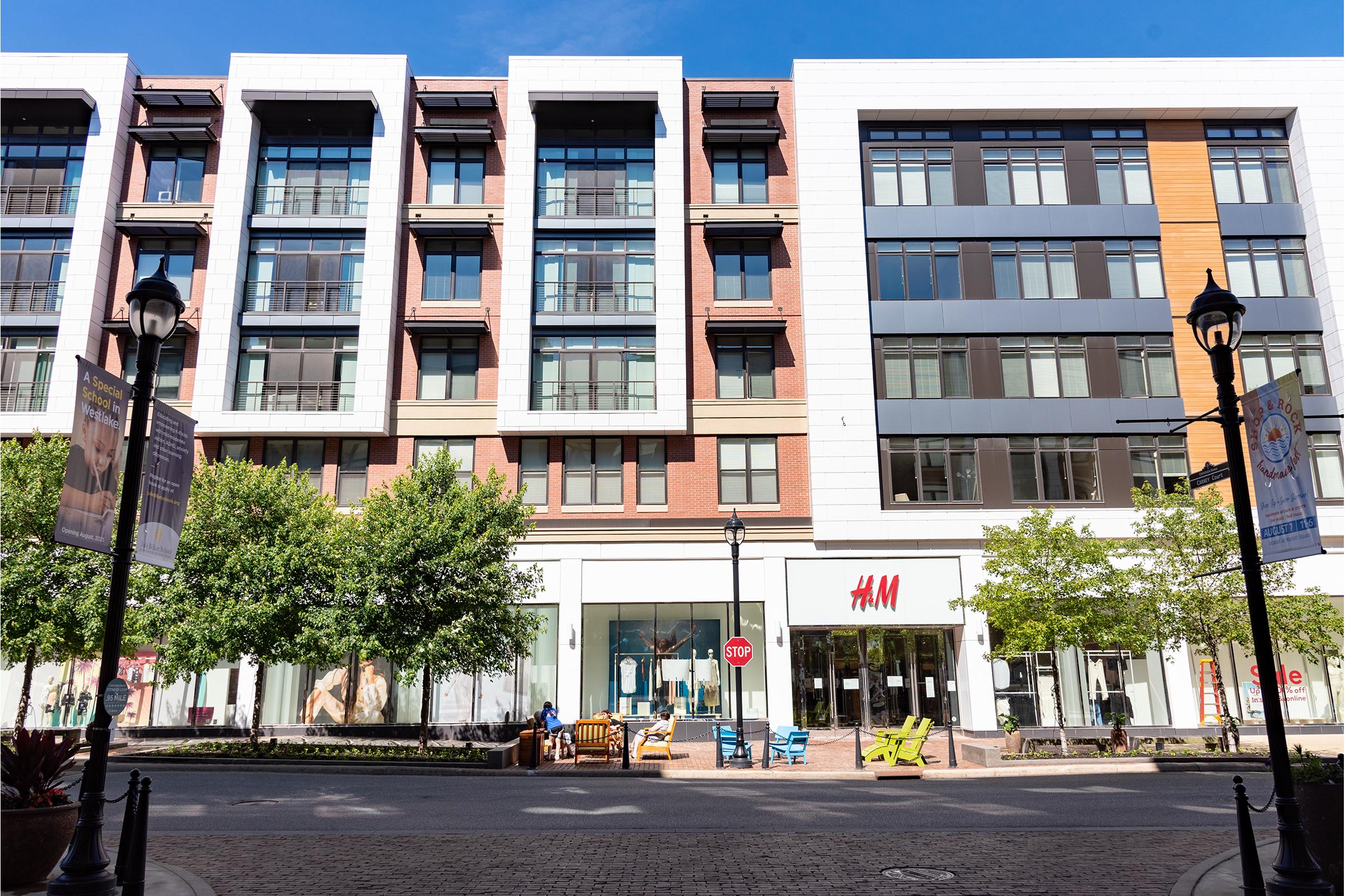 A modern urban building showcasing a storefront marked with the H&M logo. The structure features a mix of brick and glass, with trees and outdoor seating in front. Clear blue sky above adds to the vibrant atmosphere of the shopping area.