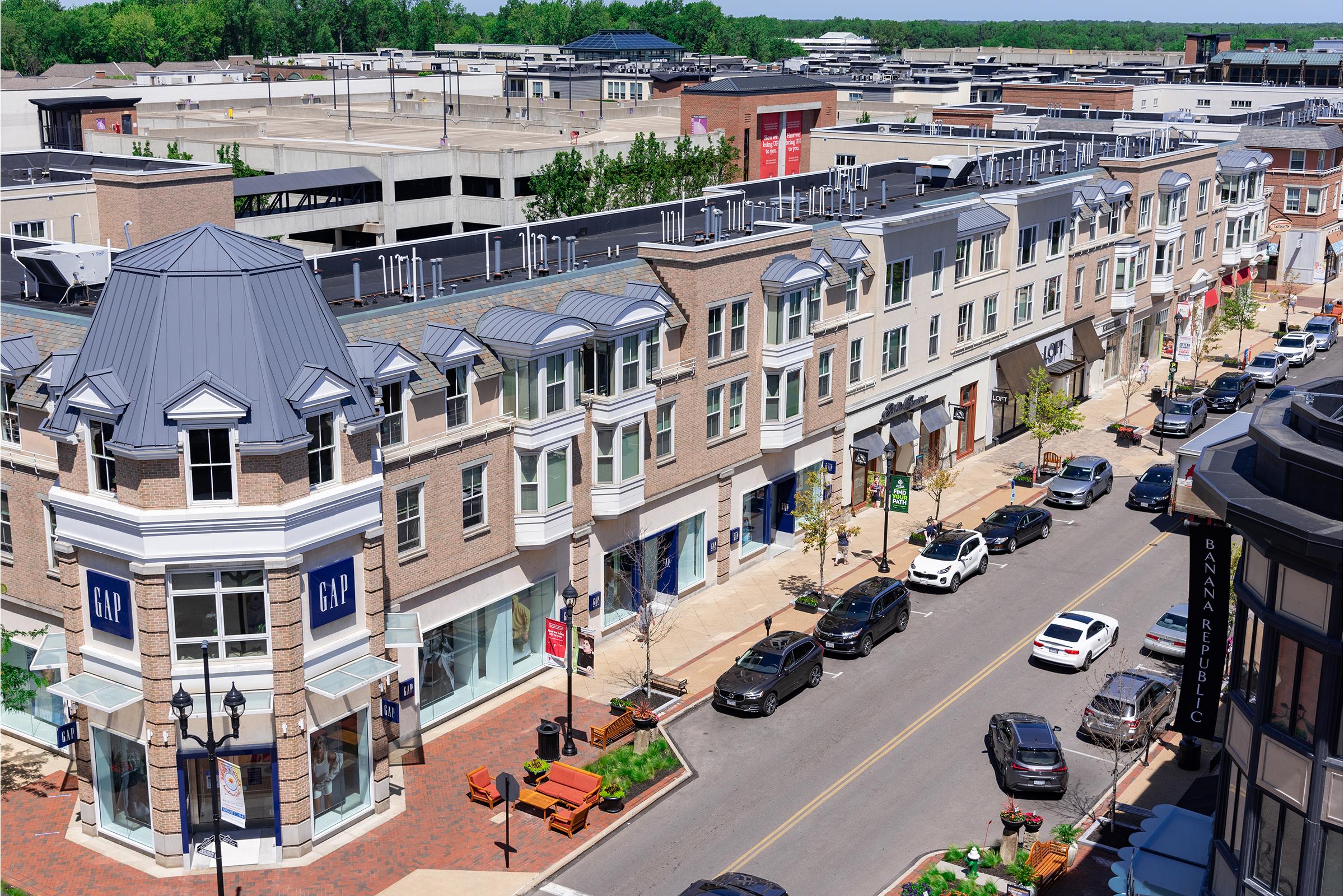 Aerial view of a shopping area featuring brick buildings with retail stores, including a GAP storefront. The street is lined with parked cars and benches, surrounded by greenery. The scene conveys a vibrant, bustling commercial atmosphere.