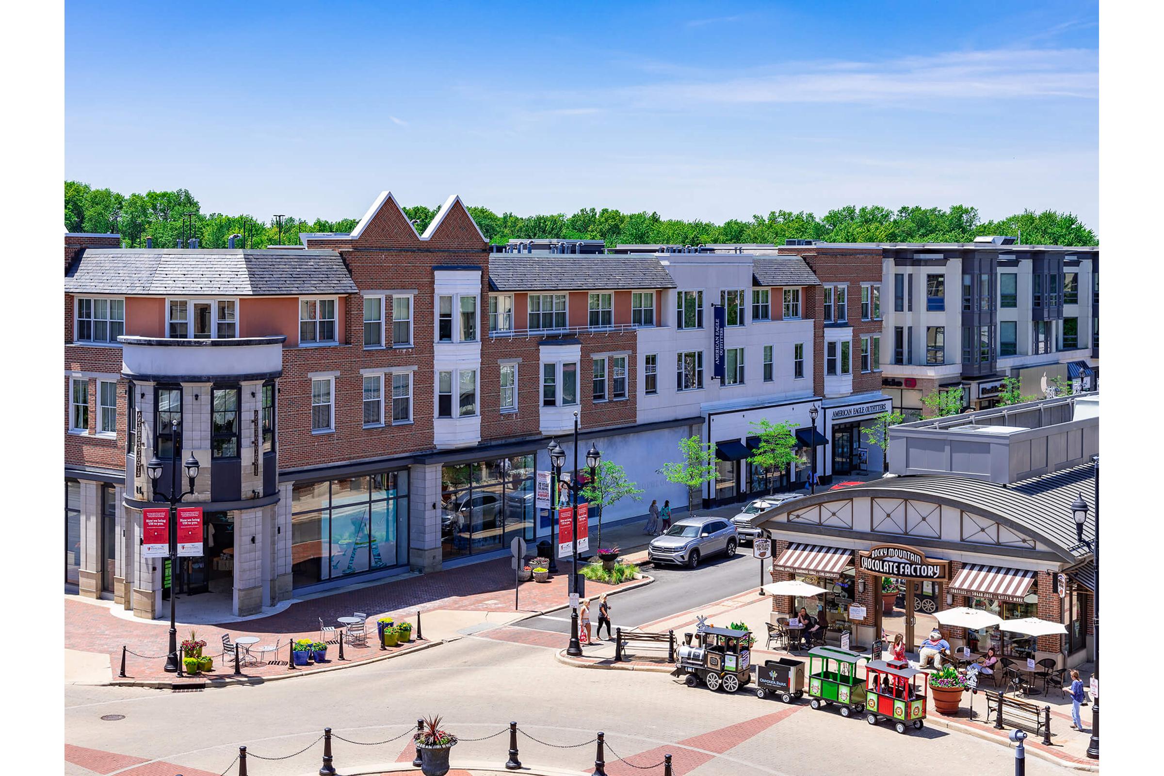 A vibrant urban scene featuring a mix of modern and traditional buildings, a bustling street filled with cars, and a charming outdoor market area. Bright blue skies and lush green trees in the background add to the lively atmosphere.