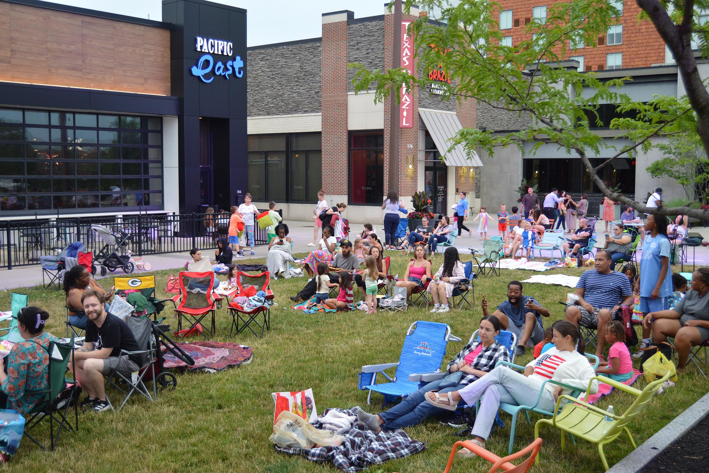 A lively outdoor scene at a community event with people sitting on lawn chairs and blankets. Families and friends gather on the grass in front of a building, enjoying the atmosphere. Some are chatting, while others are engaged in activities, with colorful decorations and a mix of clothing styles visible.