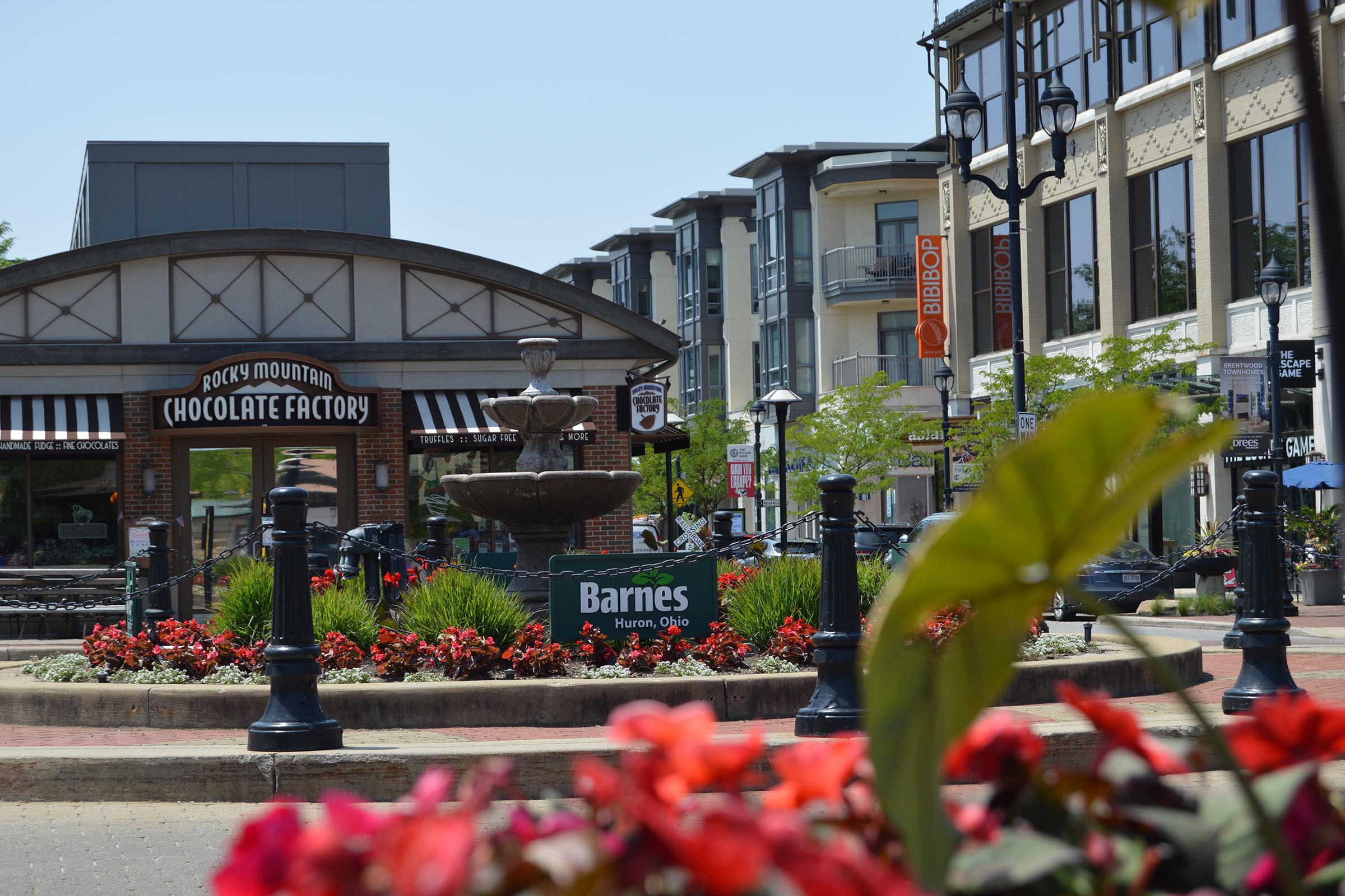 A vibrant outdoor scene featuring a fountain at a plaza, with colorful flowers in the foreground. The Rocky Mountain Chocolate Factory is visible under a striped awning. Nearby storefronts, including a sign for Barnes and several other shops, line the street, surrounded by trees and decorative street lamps.