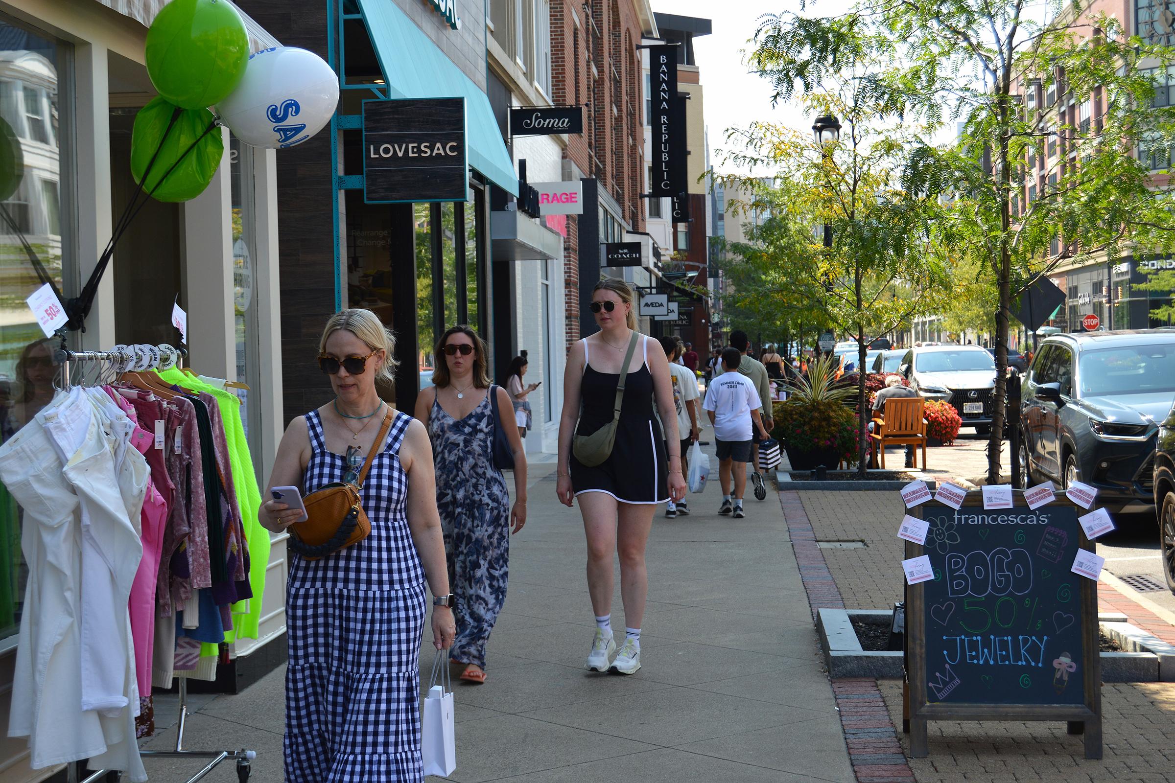 People walking along a sunny street lined with shops. A woman in a checkered dress uses her phone, while two others stroll nearby. Balloons and shop signs are visible, and a sign advertising jewelry is on a nearby sidewalk. Trees provide greenery to the urban scene.