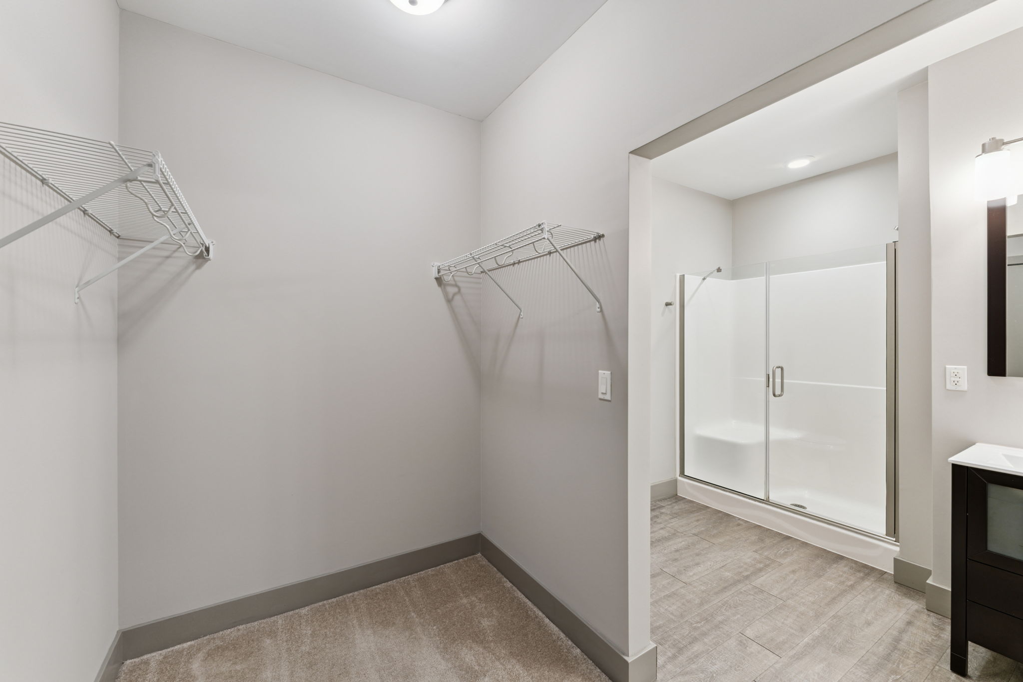 Interior view of a bathroom featuring a clear glass shower, a dark cabinet with a mirror, and two wire shelving units on the wall. The walls are painted in a neutral color, and the floor has a light tile design, creating a modern and spacious atmosphere.